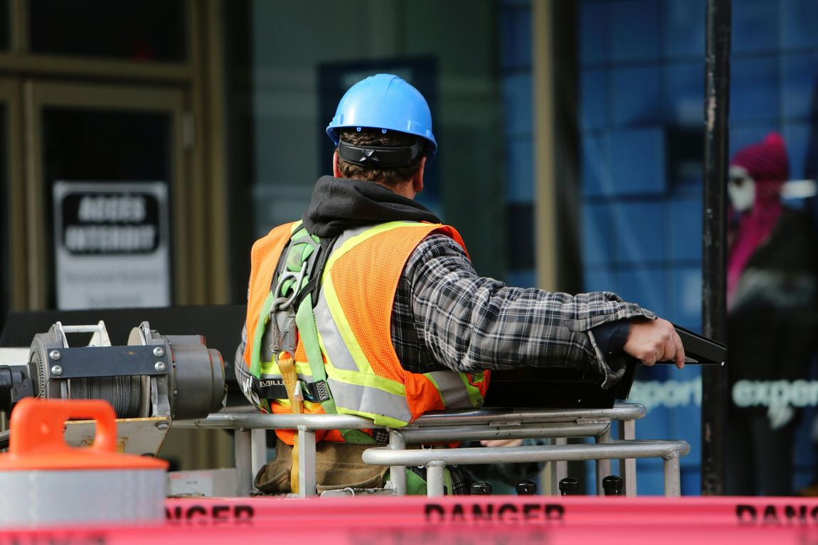 Ouvrier du bâtiment portant un casque bleu, un gilet orange et un harnais de sécurité, utilisant un équipement à proximité d'un bâtiment avec du ruban de précaution.