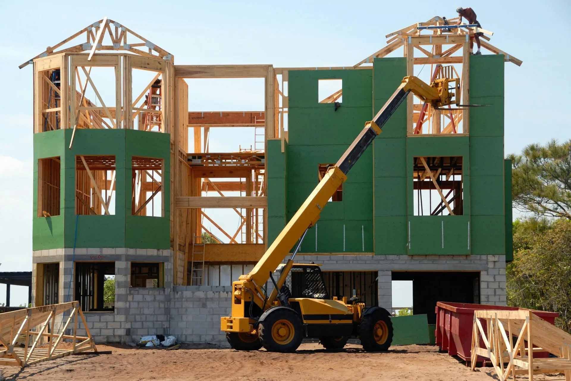 Chantier : charpente de maison avec revêtement vert, chariot élévateur jaune et ciel bleu.