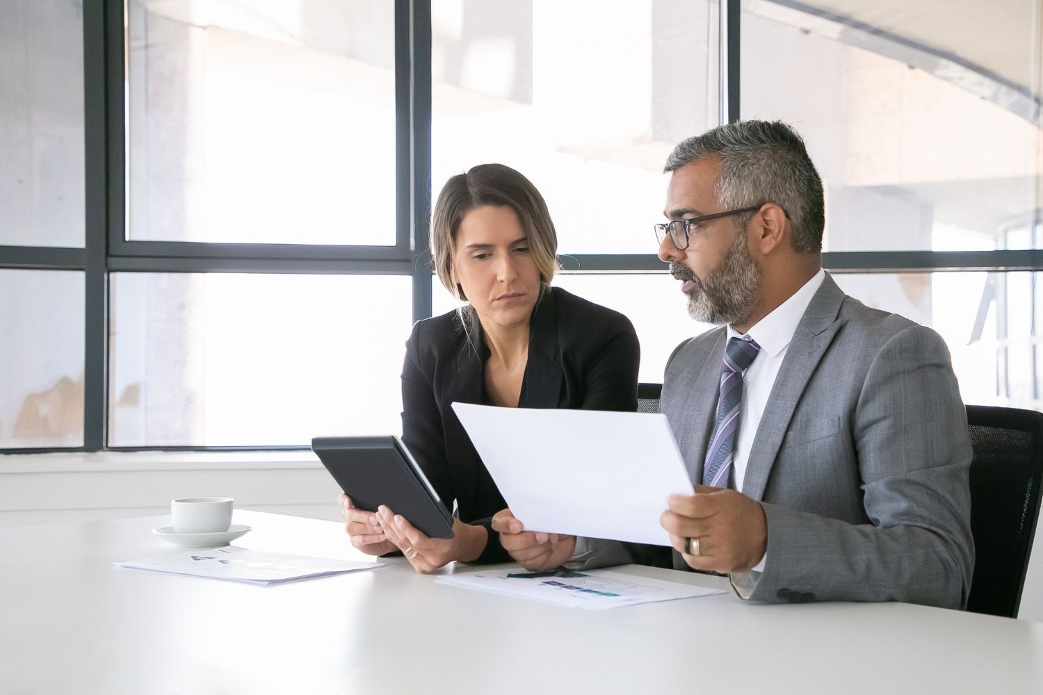 Man and woman in suits reviewing documents at a table, near a window.