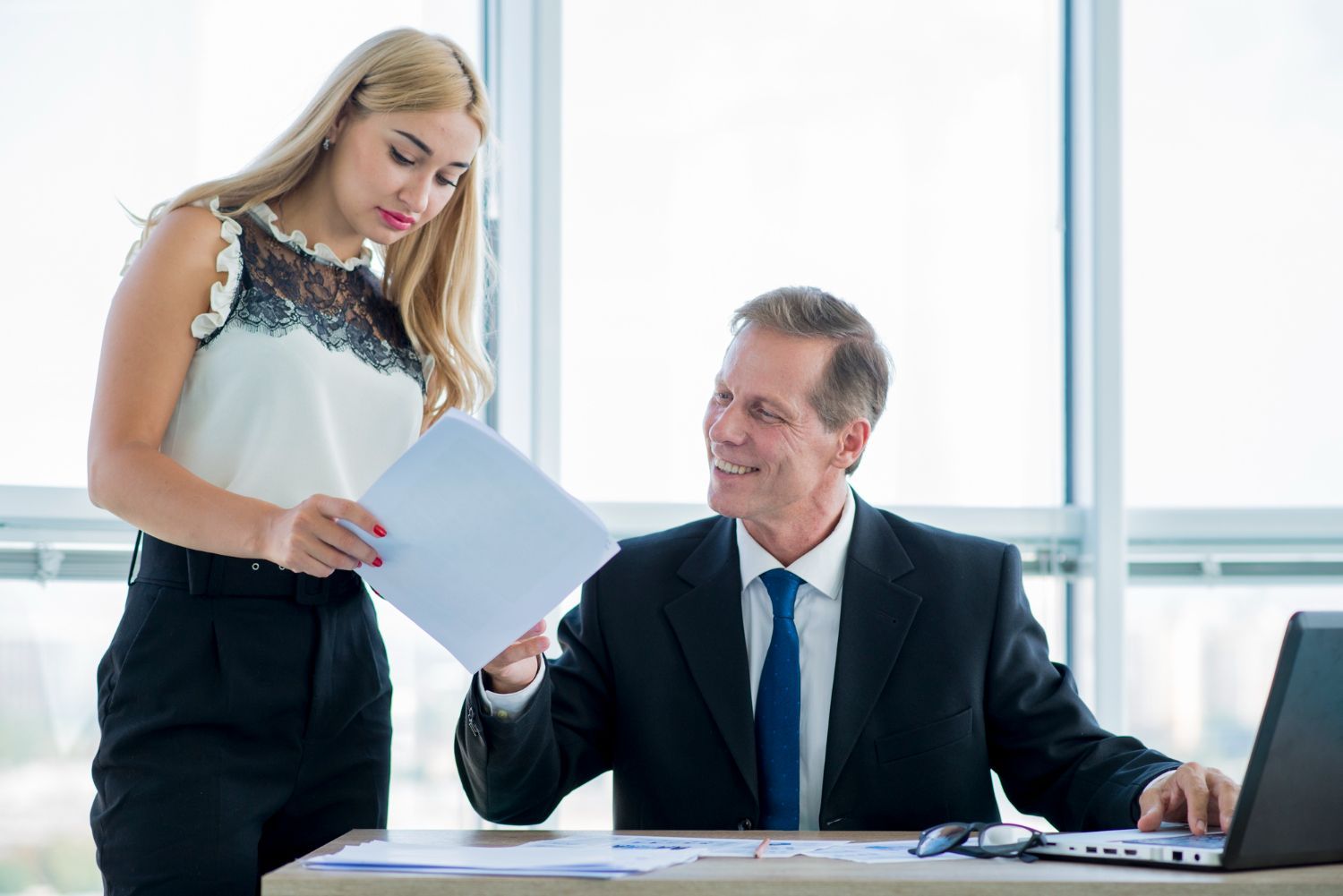 Woman handing papers to a man in an office setting. They both appear to be smiling.