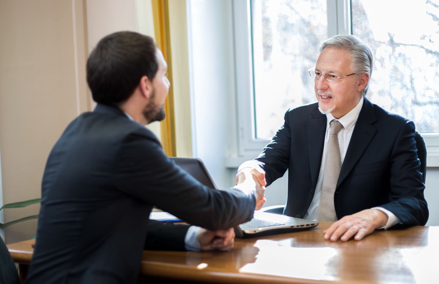 Two men in suits shaking hands at a table, likely in an office setting.
