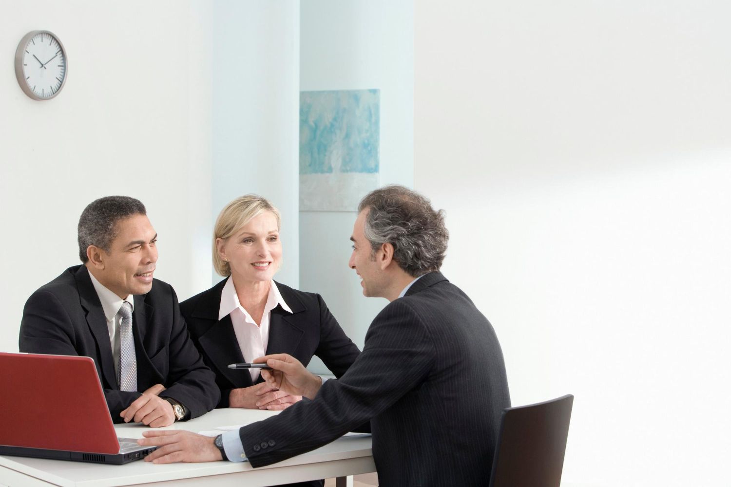Three people in suits at a table; one points, others smile. Laptop and clock in the background.