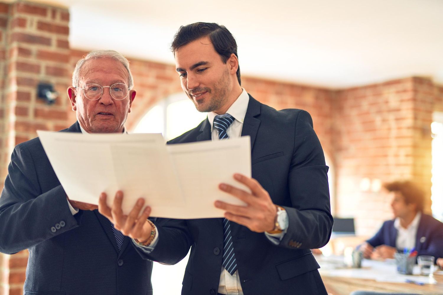 Two men in suits reviewing papers, smiling, indoors.