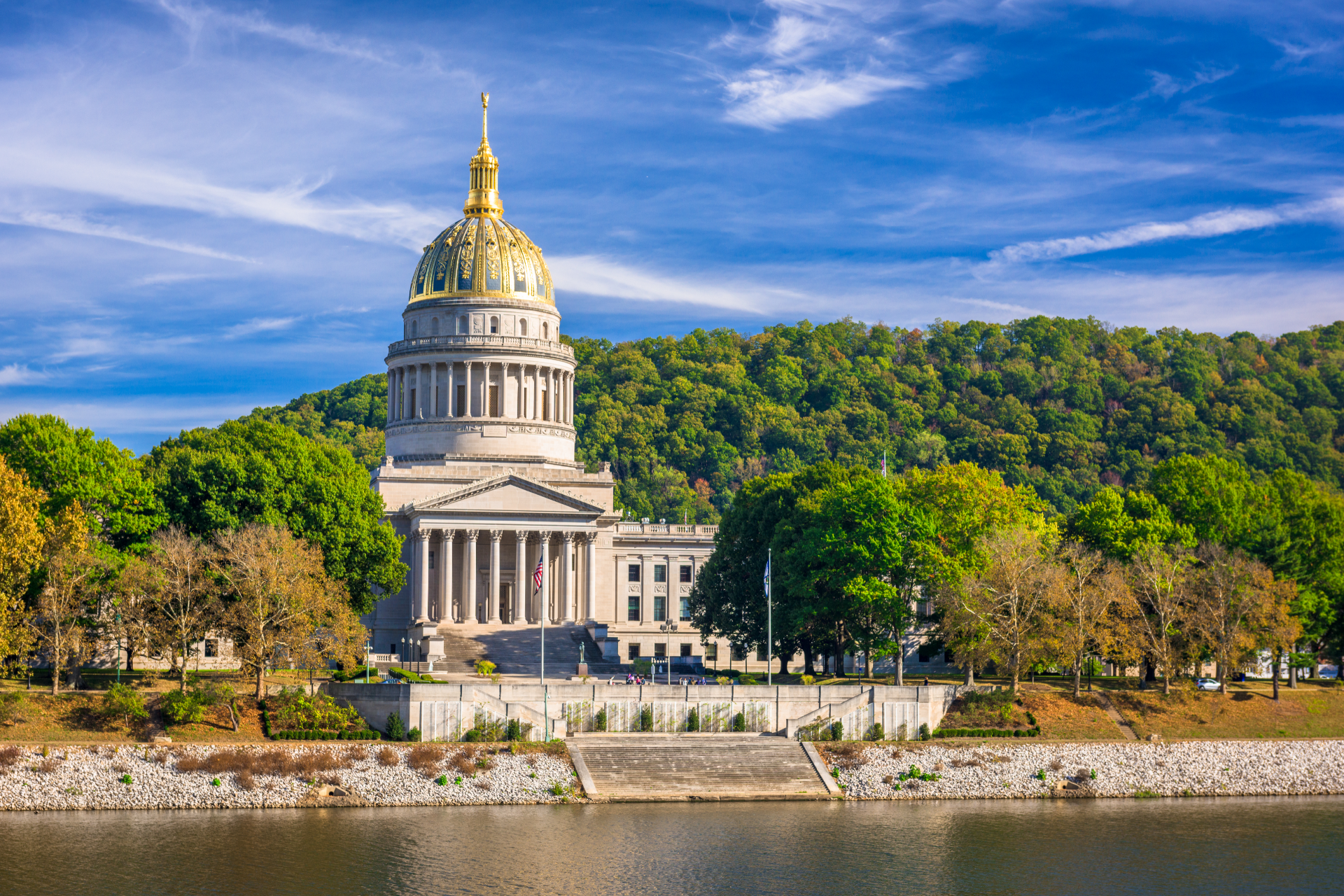 West Virginia State Capitol building with golden dome, overlooking river and trees on sunny day.