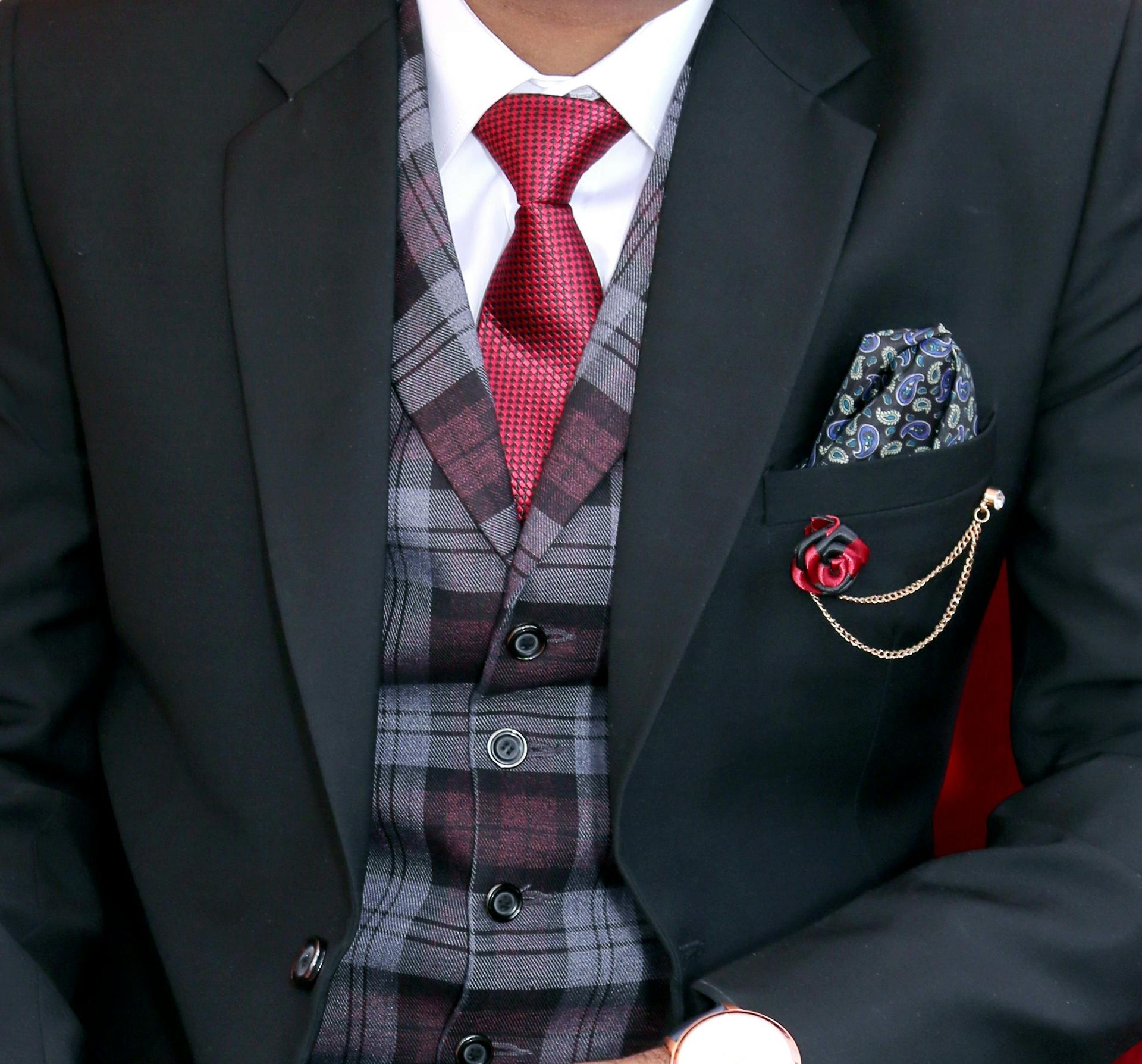 Man in a black suit, plaid vest, and red tie with a floral lapel pin and pocket square.