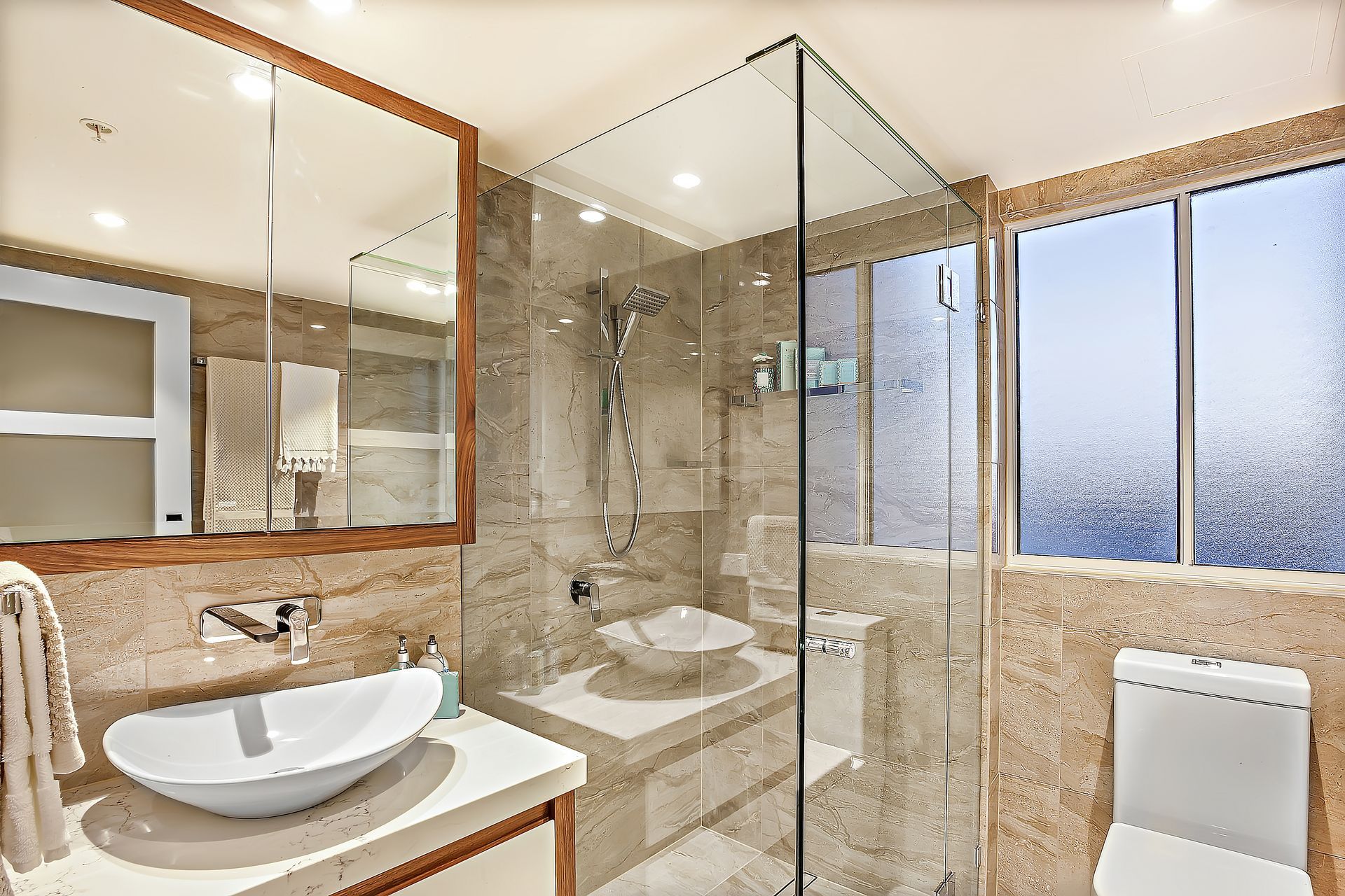 Modern washroom, shower area and washing area, including a white sink and silver taps on the ceramic counter beside glass cover and the toilet bowl, the mirror is attached to the wall