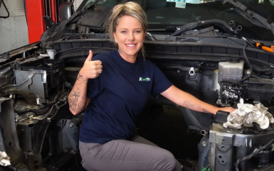 A mechanic in a navy t-shirt gives a thumbs-up while working inside the engine bay of a vehicle at an auto shop.