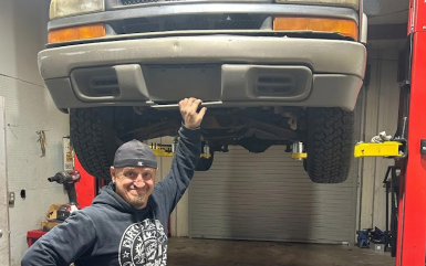 A person wearing a dark hoodie and cap smiles while holding up the front bumper of a truck raised on a mechanic's lift.