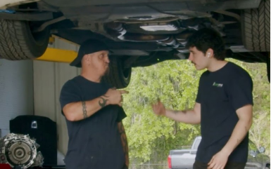 Two mechanics in black t-shirts talk beneath a car raised on a lift in a garage.