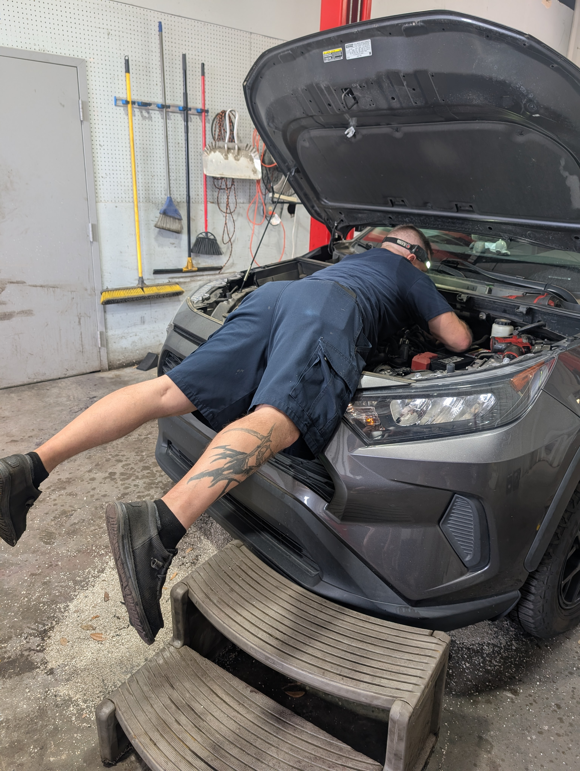 A mechanic leans over the open hood of a gray car in a repair shop, using a step stool to reach the engine.