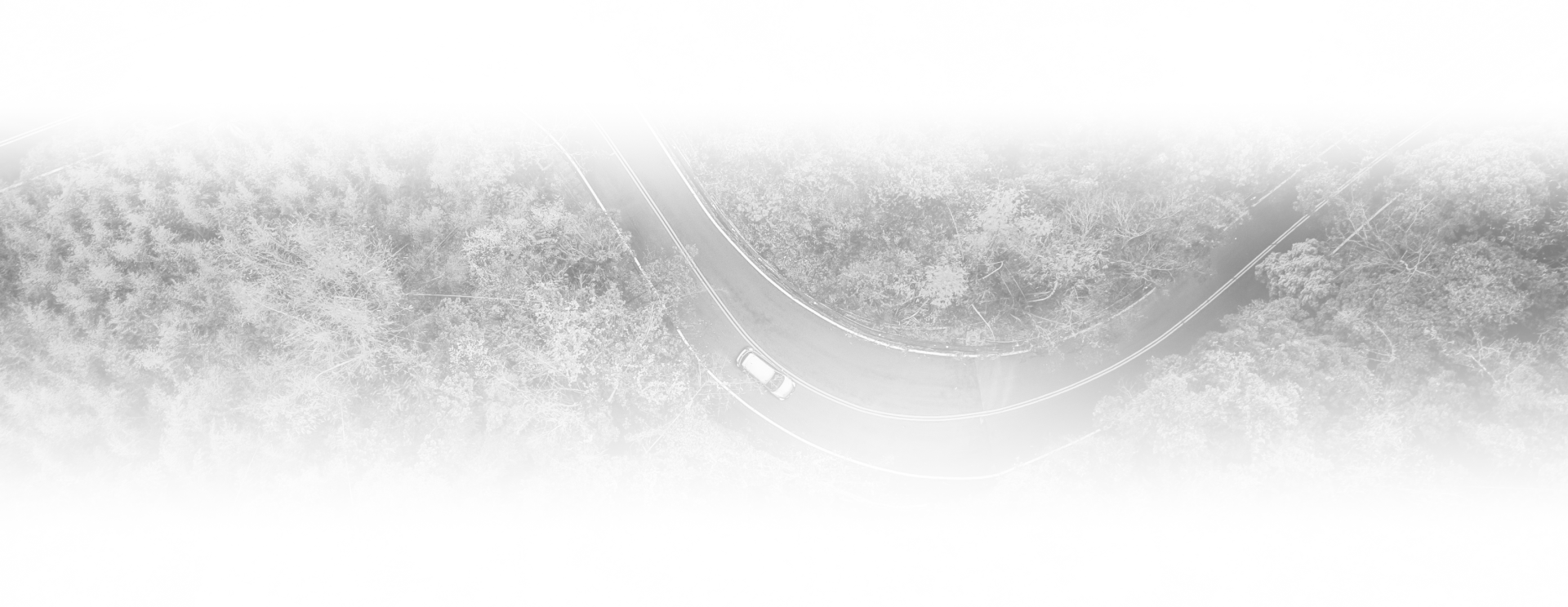 An aerial view of a single white car driving along a curved road through a dense, foggy forest.