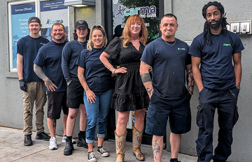 A group of seven people in dark blue work uniforms stand in a line outside a Carma Auto Grill building entrance.