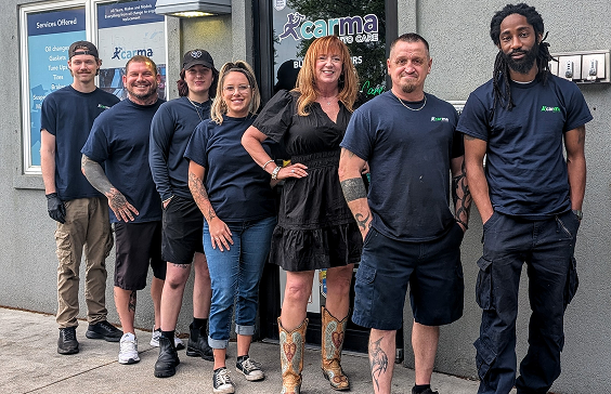 A group of seven people in dark blue work uniforms stand in a line outside a Carma Auto Grill building entrance.