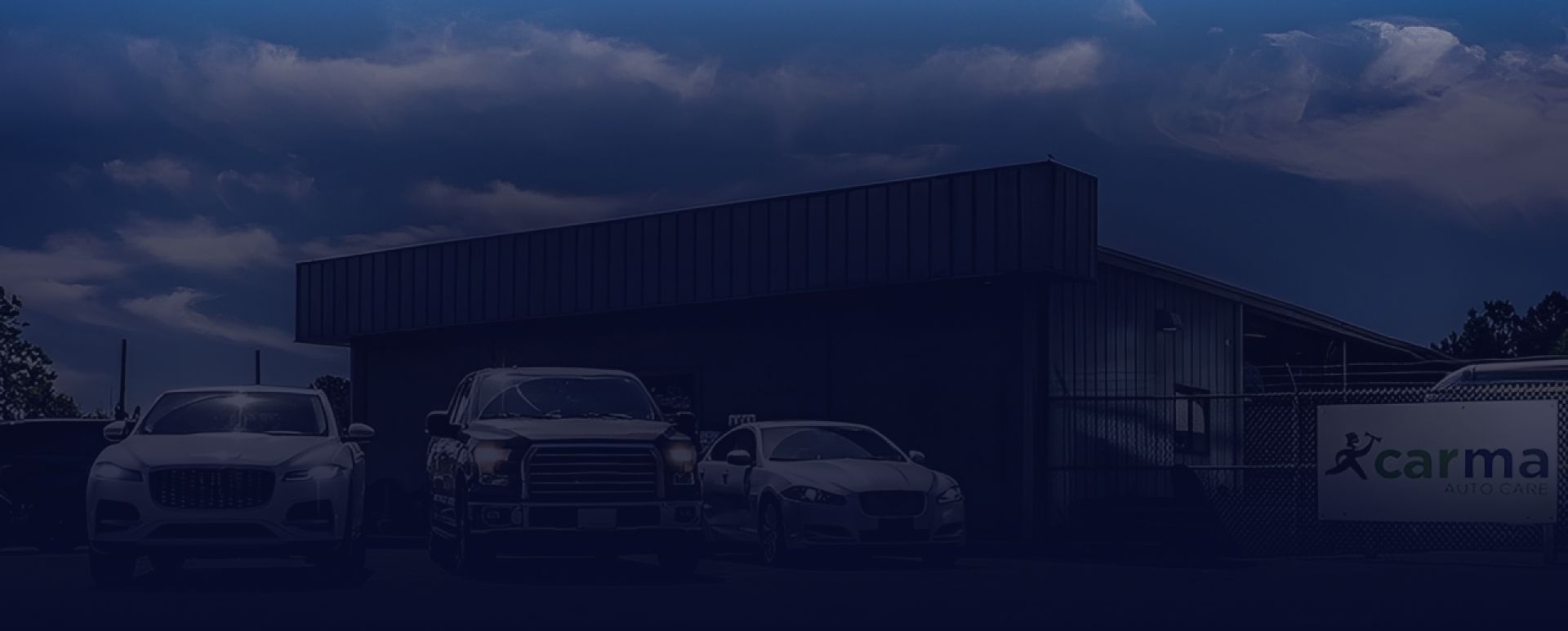 Three white vehicles parked in front of a commercial building with a Carma sign under a cloudy sky | CARma Auto Care