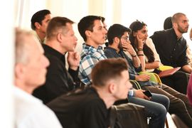An audience dressed in professional attire sit in a row of chairs, all looking in the same direction attentively, as if listening to a speaker.
