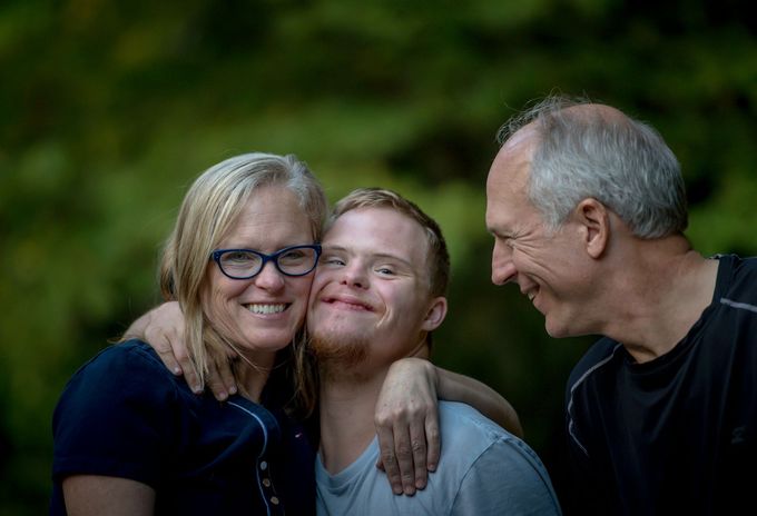 Two parents sit on either side of a teenager with Down Syndrome with one arm around each of their shoulders. The teen is smiling and pressing their cheek up against the parent's cheek.