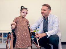 A medical professional in a white coat crouches down beside a small child in a flowered dress and glasses as she supports herself using a mobility device.
