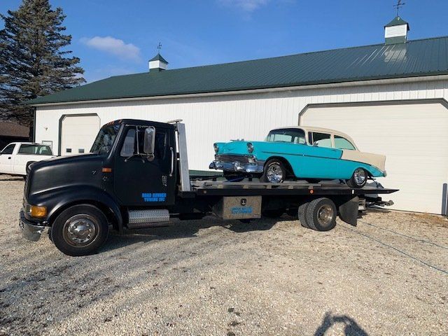 A tow truck with two cars on the back is parked in front of a garage.