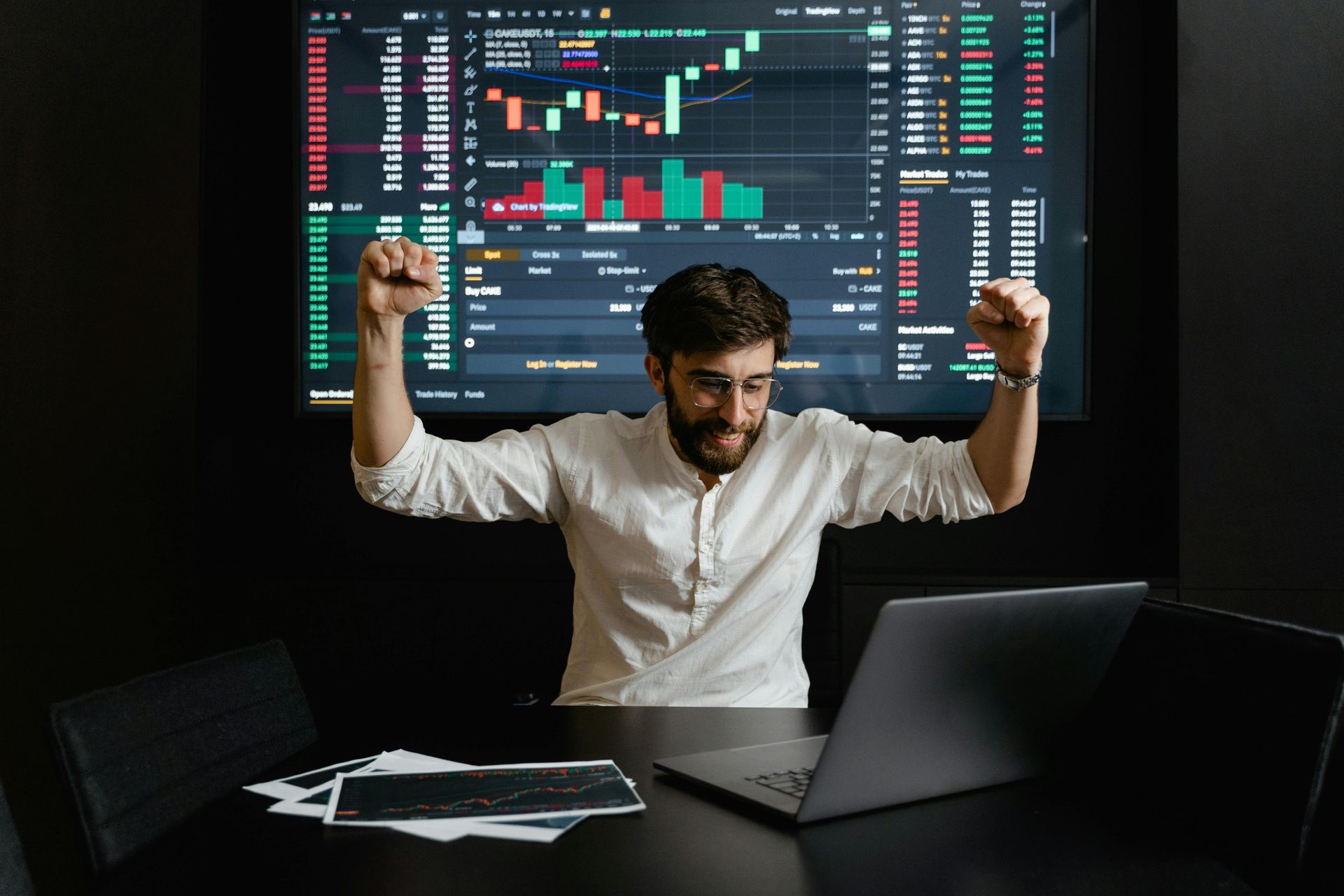 A man celebrates in front of data on a large screen; a laptop and papers are on the desk.