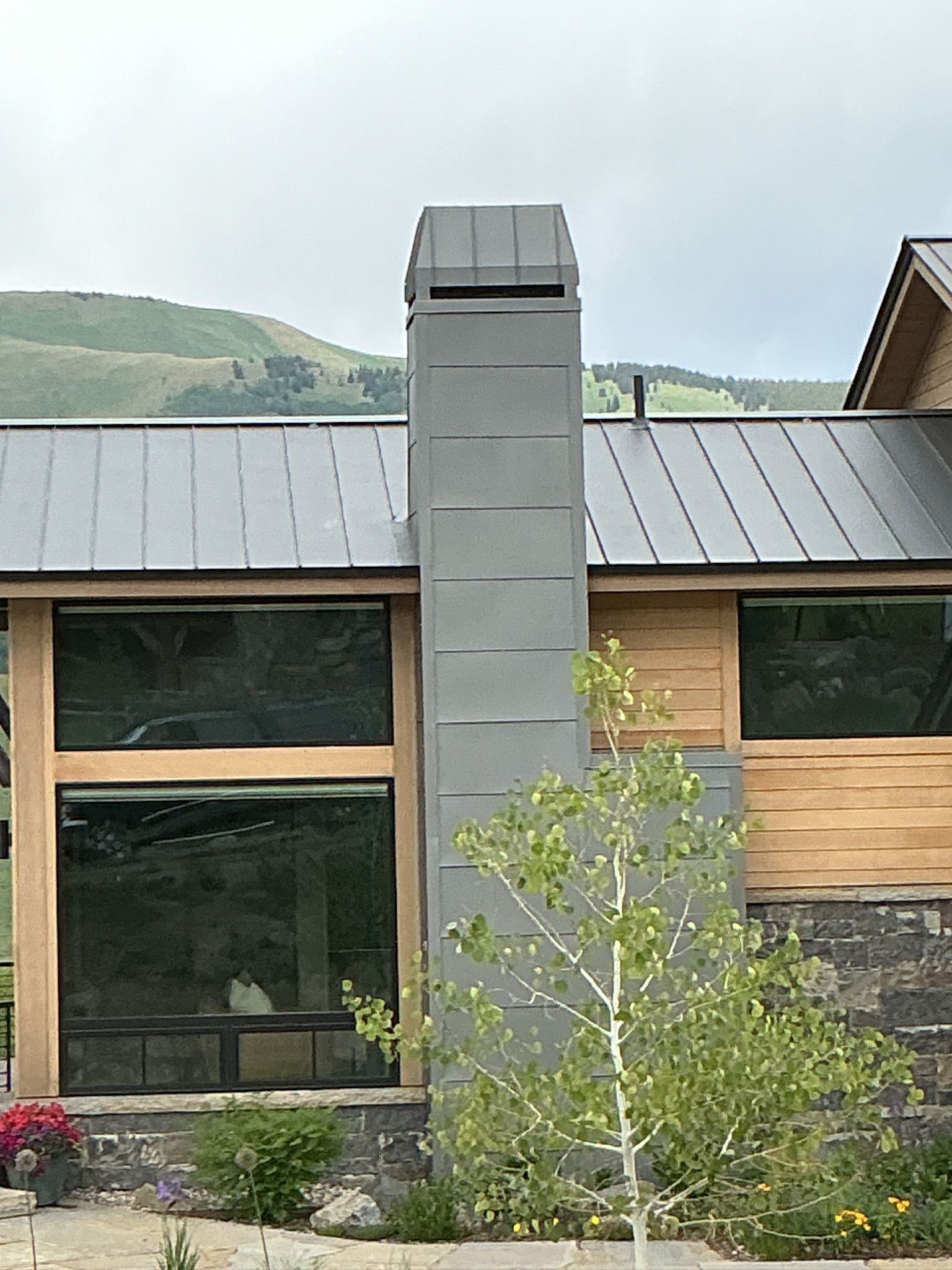Gray metal chimney on a house with a dark metal roof and large windows.