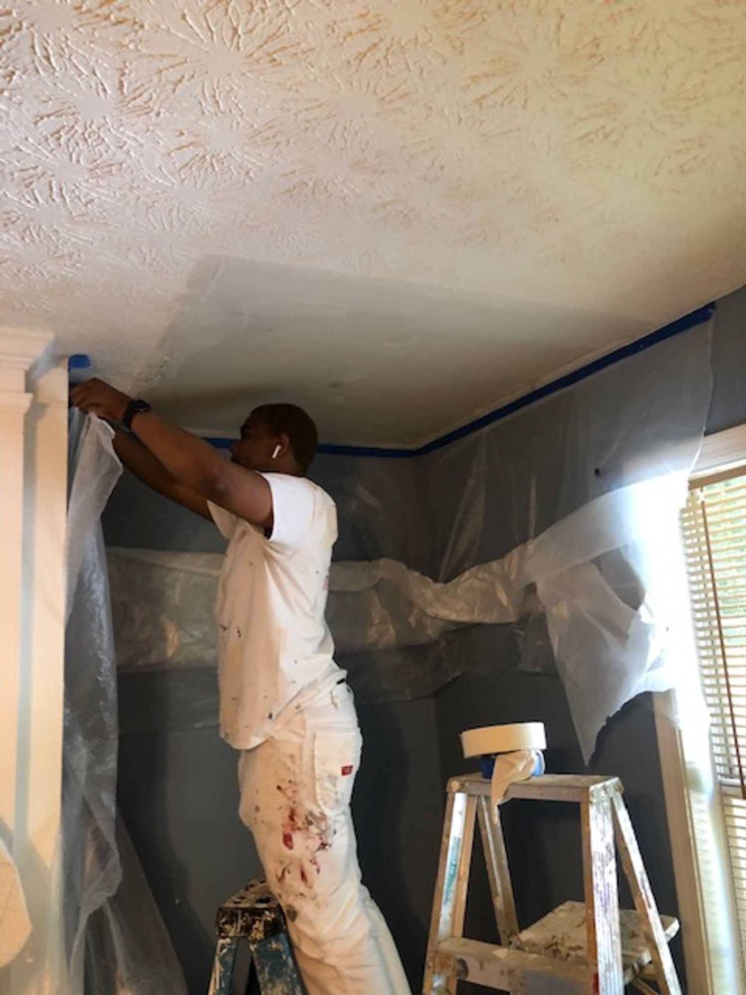 A man is standing on a ladder painting the ceiling of a room.