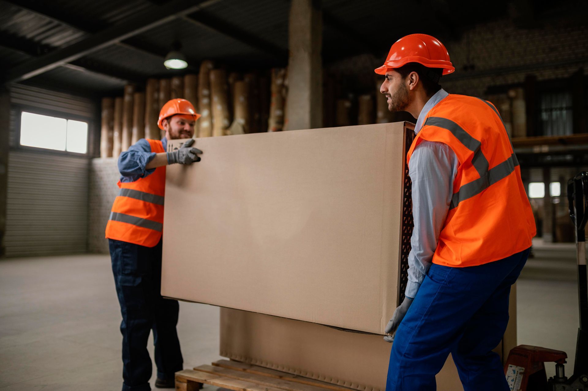 Two men are carrying a large piece of cardboard in a warehouse.