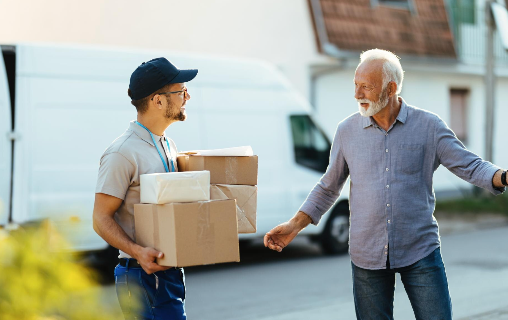 A delivery man is carrying boxes and talking to an older man.
