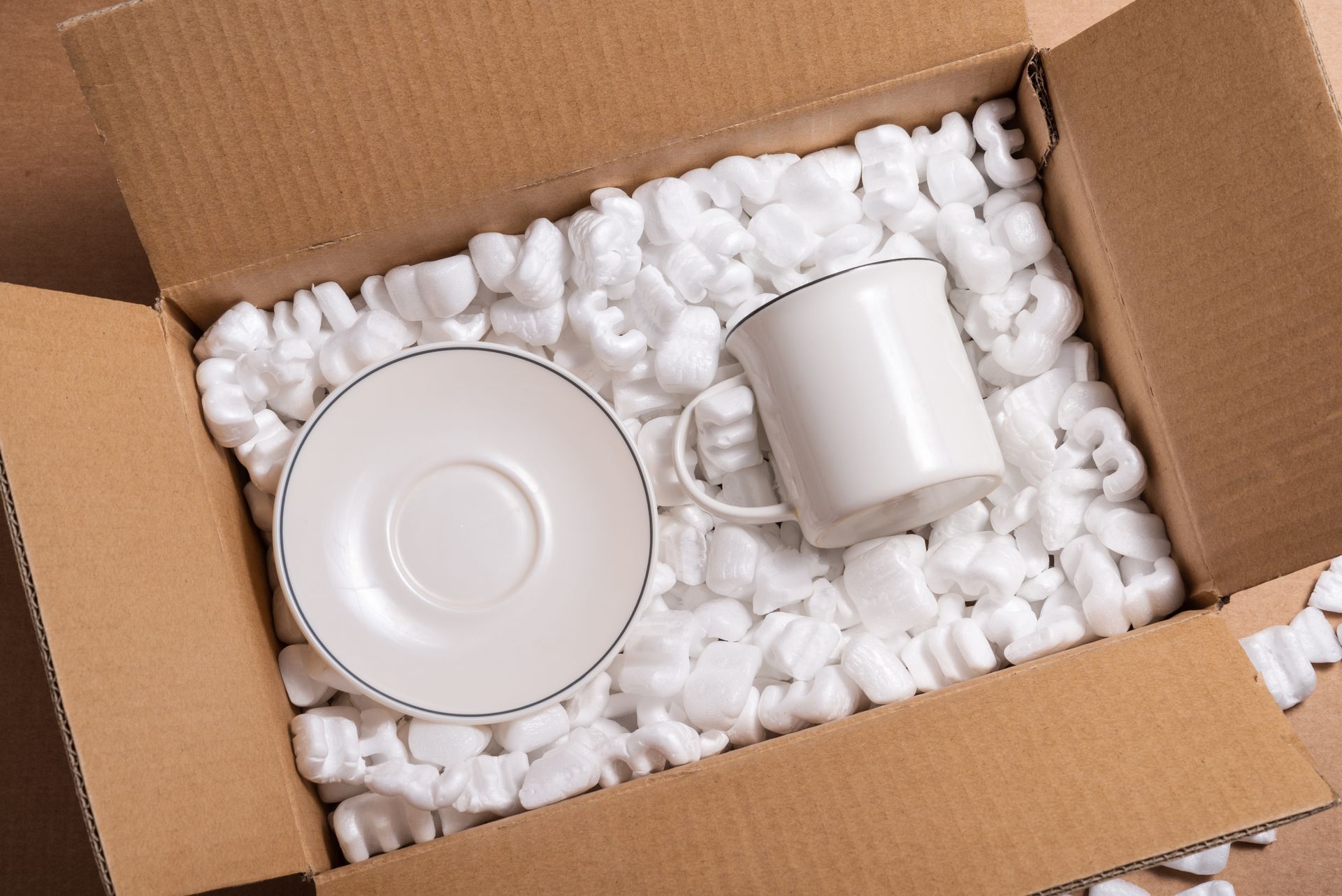 A cup and saucer are in a cardboard box filled with styrofoam peanuts.