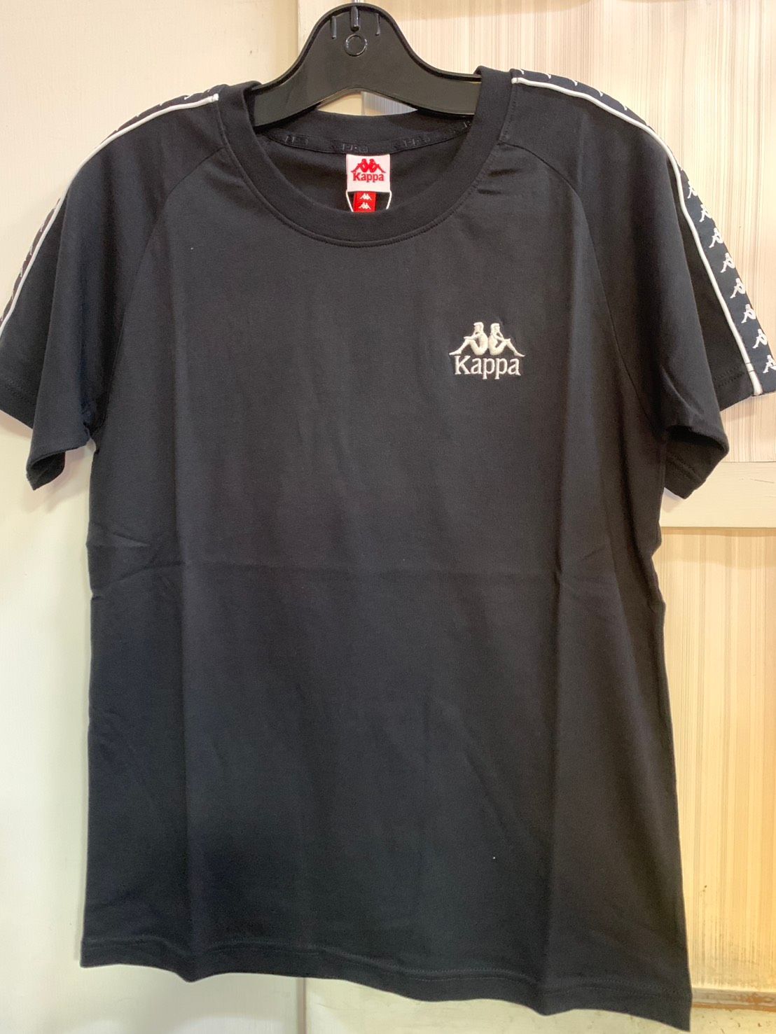 白色 Kappa T 卹，肩部有黑色標誌和條紋細節，掛在黑色衣架上。