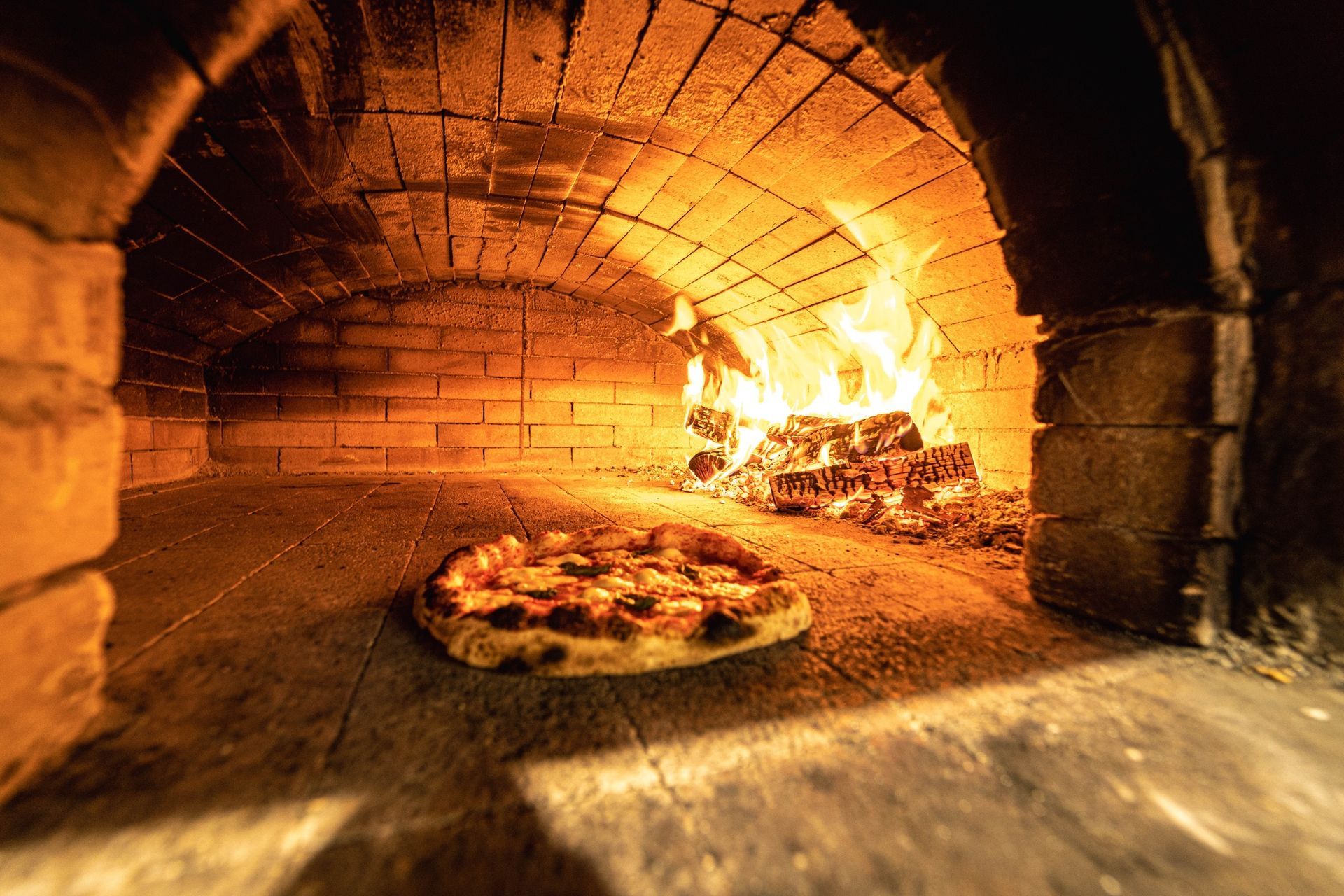 Pizza cotta in un forno a legna con fiamme sullo sfondo.