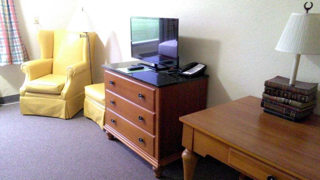 Yellow chair and ottoman next to a wooden dresser with a TV. A table with a lamp sits to the right.