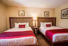 Two beds in a hotel room with red accents, a nightstand, and framed artwork.