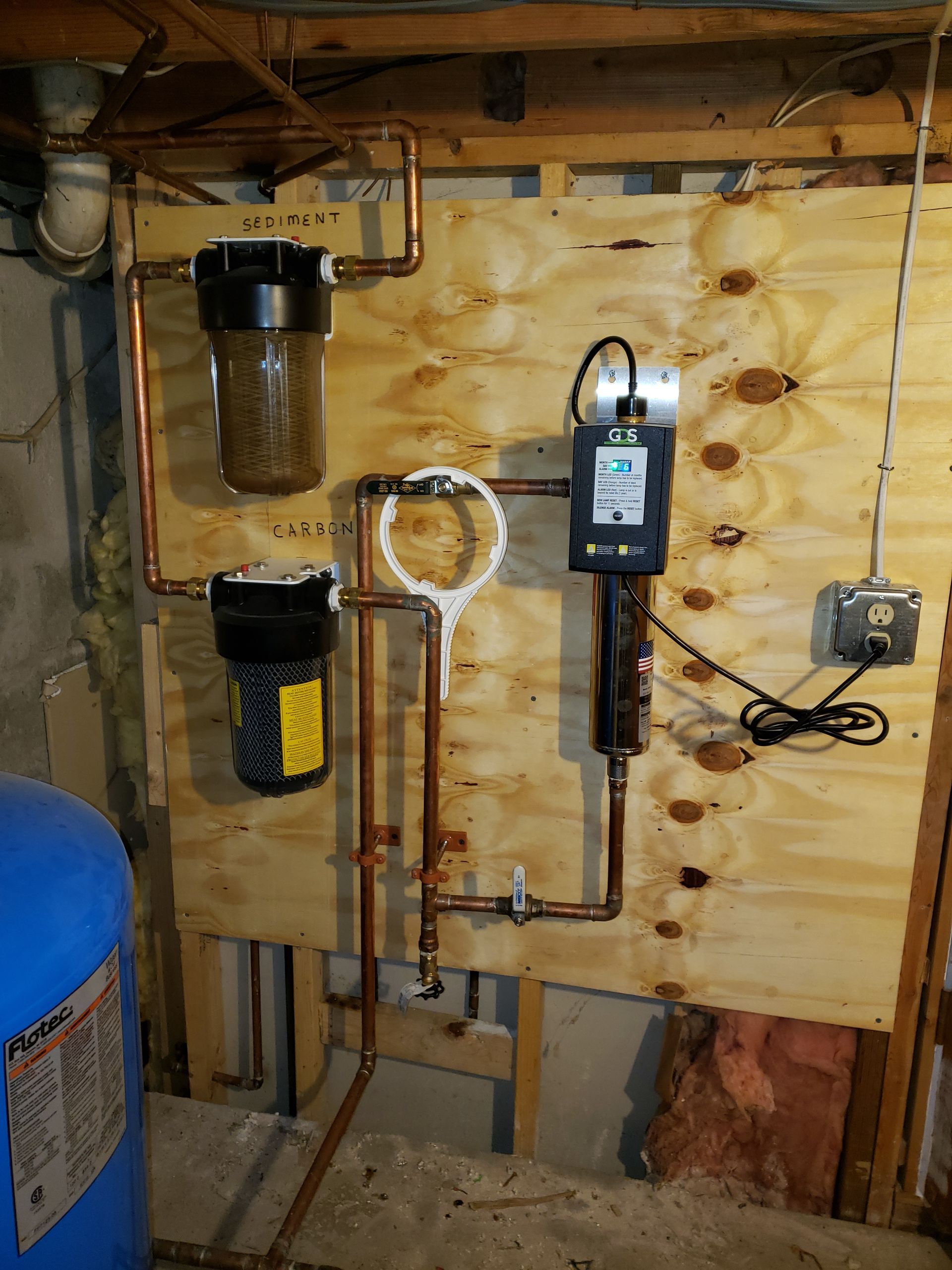 Water filtration system with copper pipes and two filter housings mounted on wood paneling.