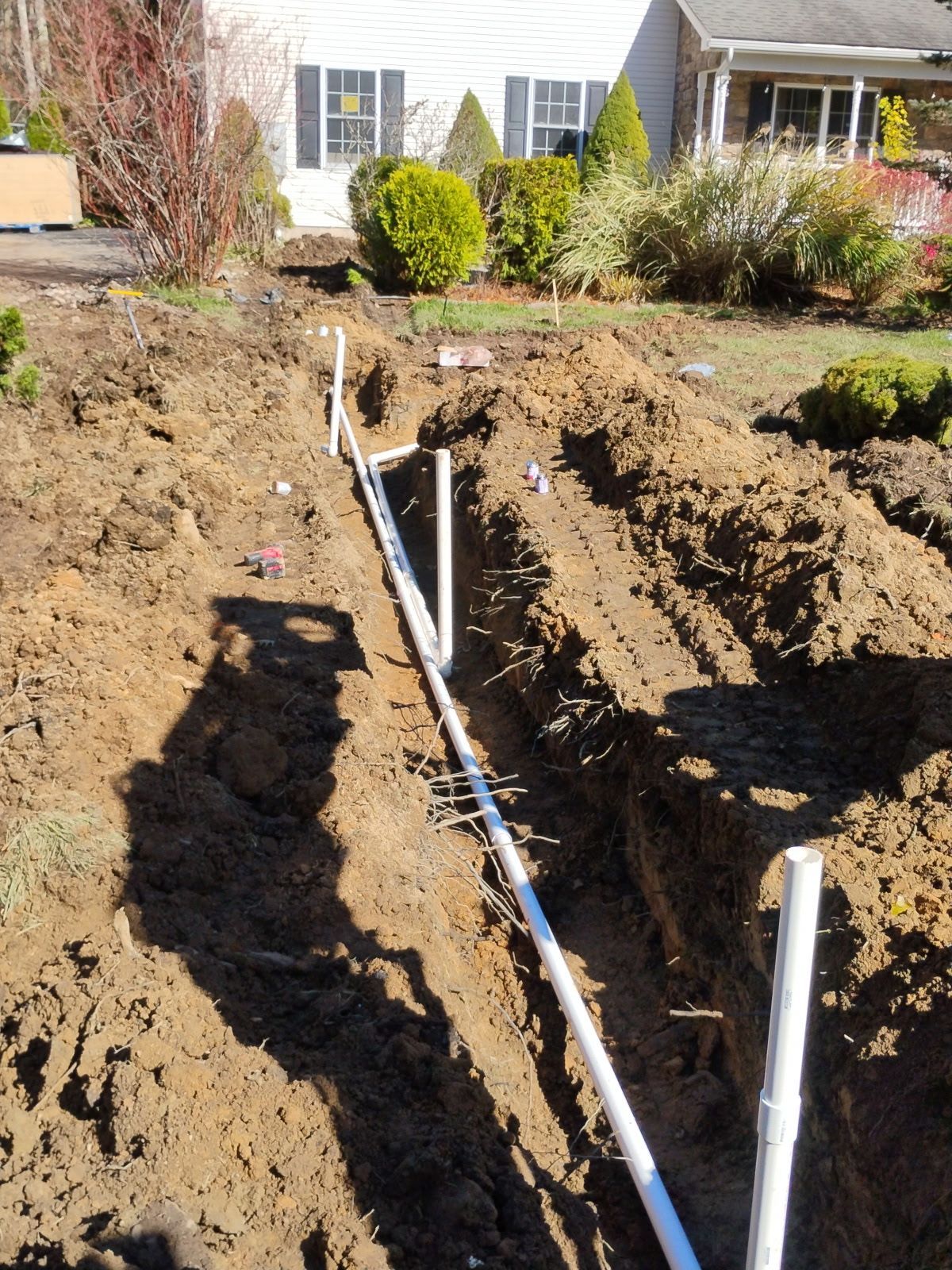 Trench in dirt with white PVC pipes laid out, likely for an irrigation system, in front of a house.