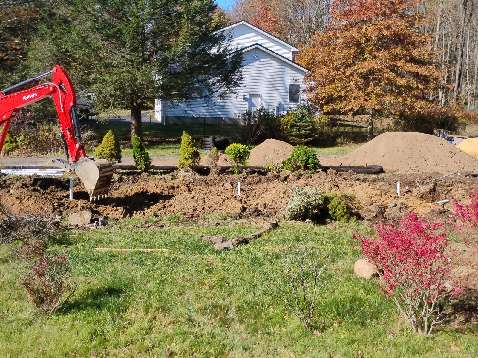 Red excavator digging in a front yard with dirt piles, near a house and trees.