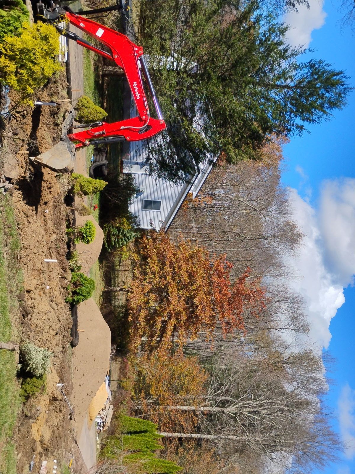 Red excavator working on landscaping near a white house and fall trees under a blue sky.