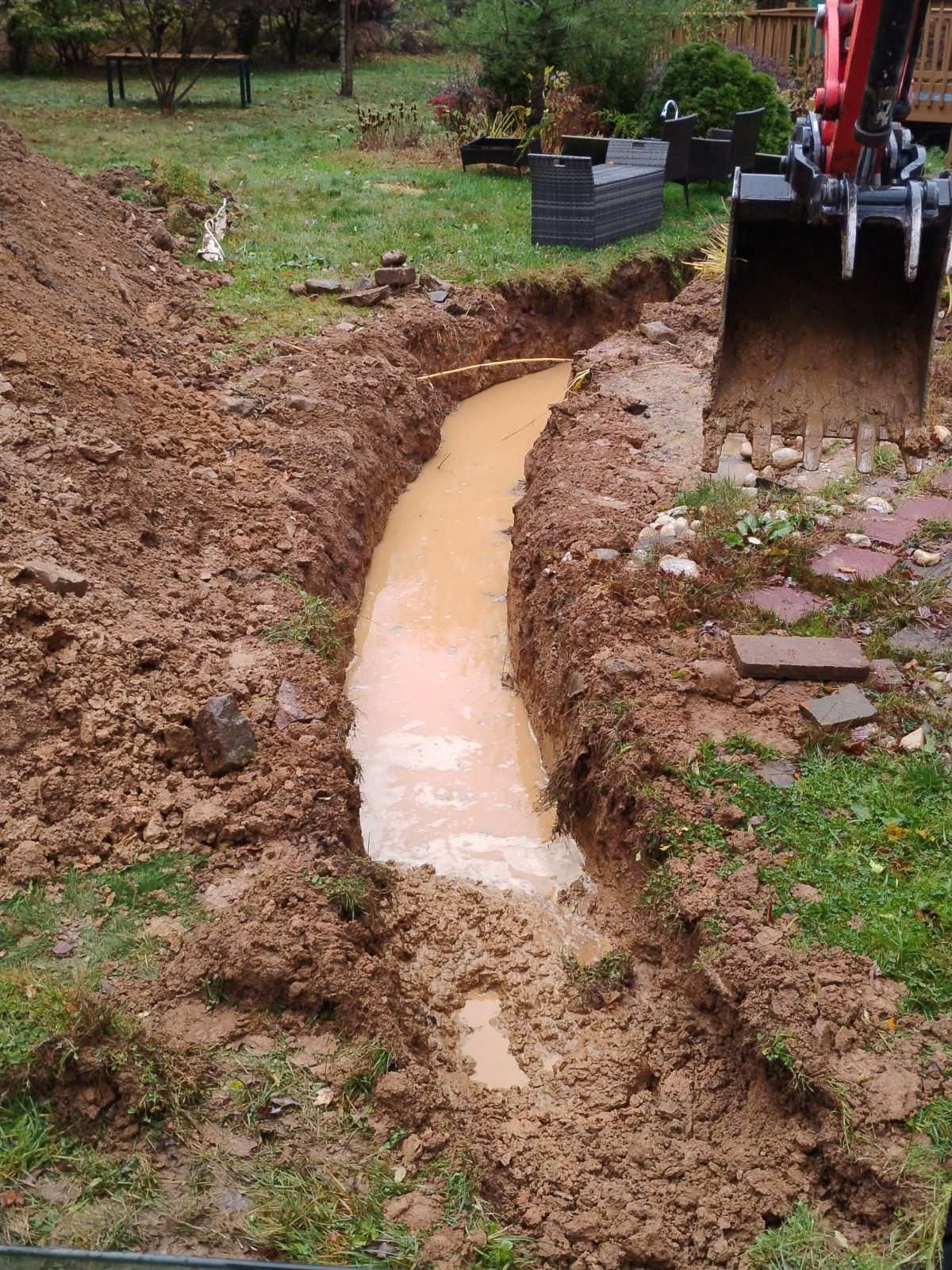A muddy trench filled with water; an excavator sits at the edge in a grassy backyard.