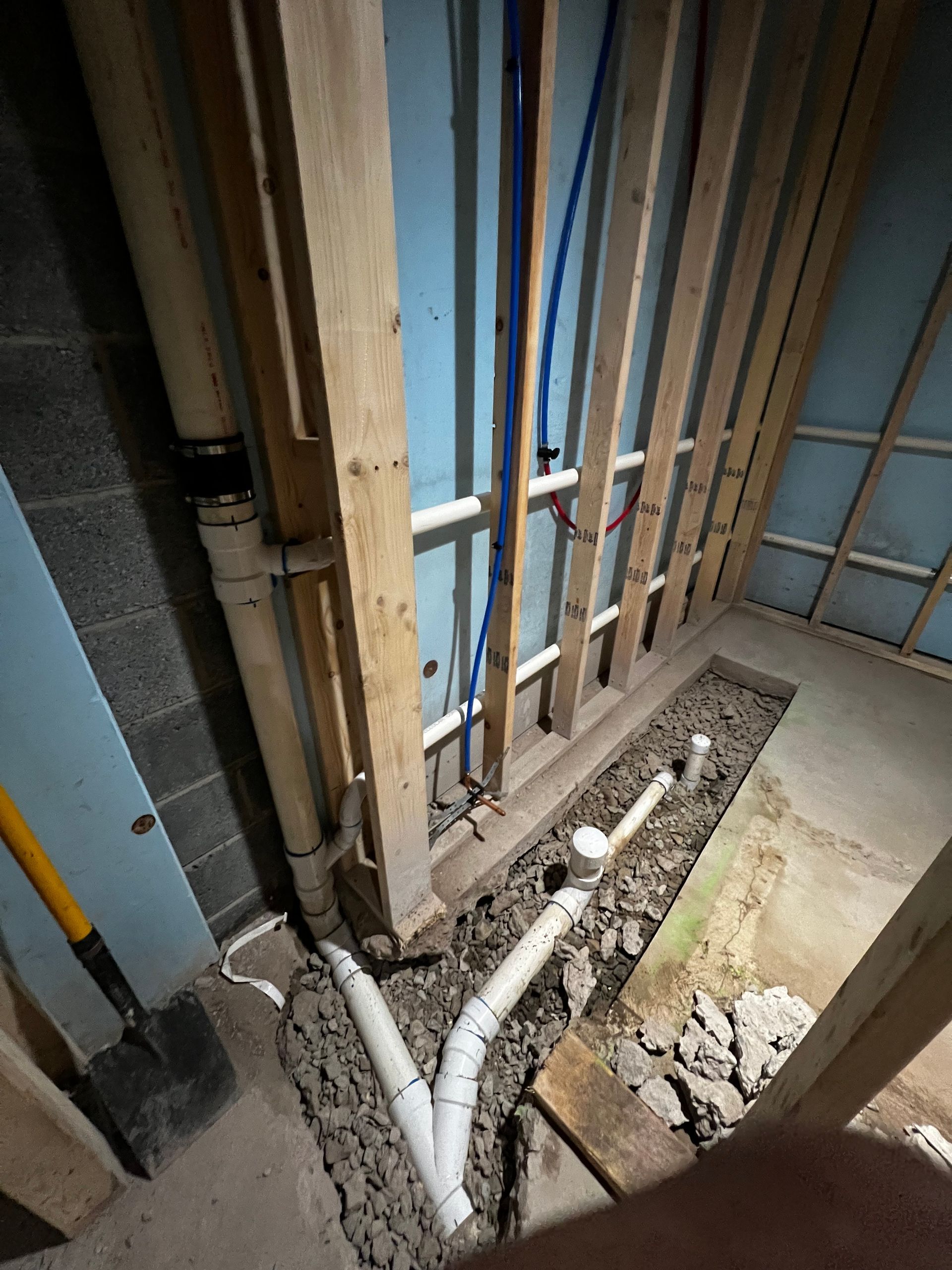 Basement wall under construction: exposed pipes, wooden framing, blue insulation, and dirt.