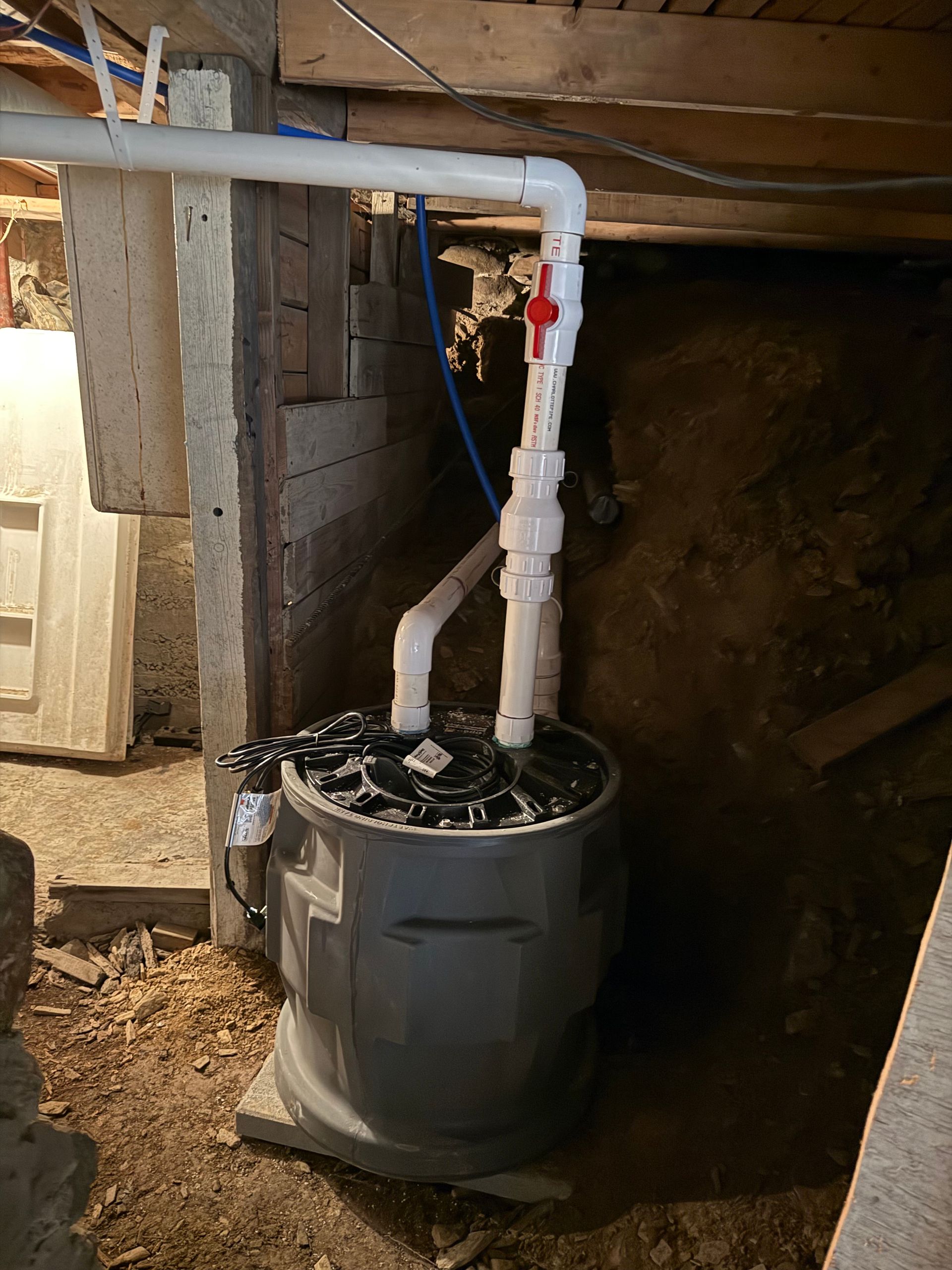 Sump pump installation in a dark basement. Gray basin, white PVC pipes, red valve, blue tube.