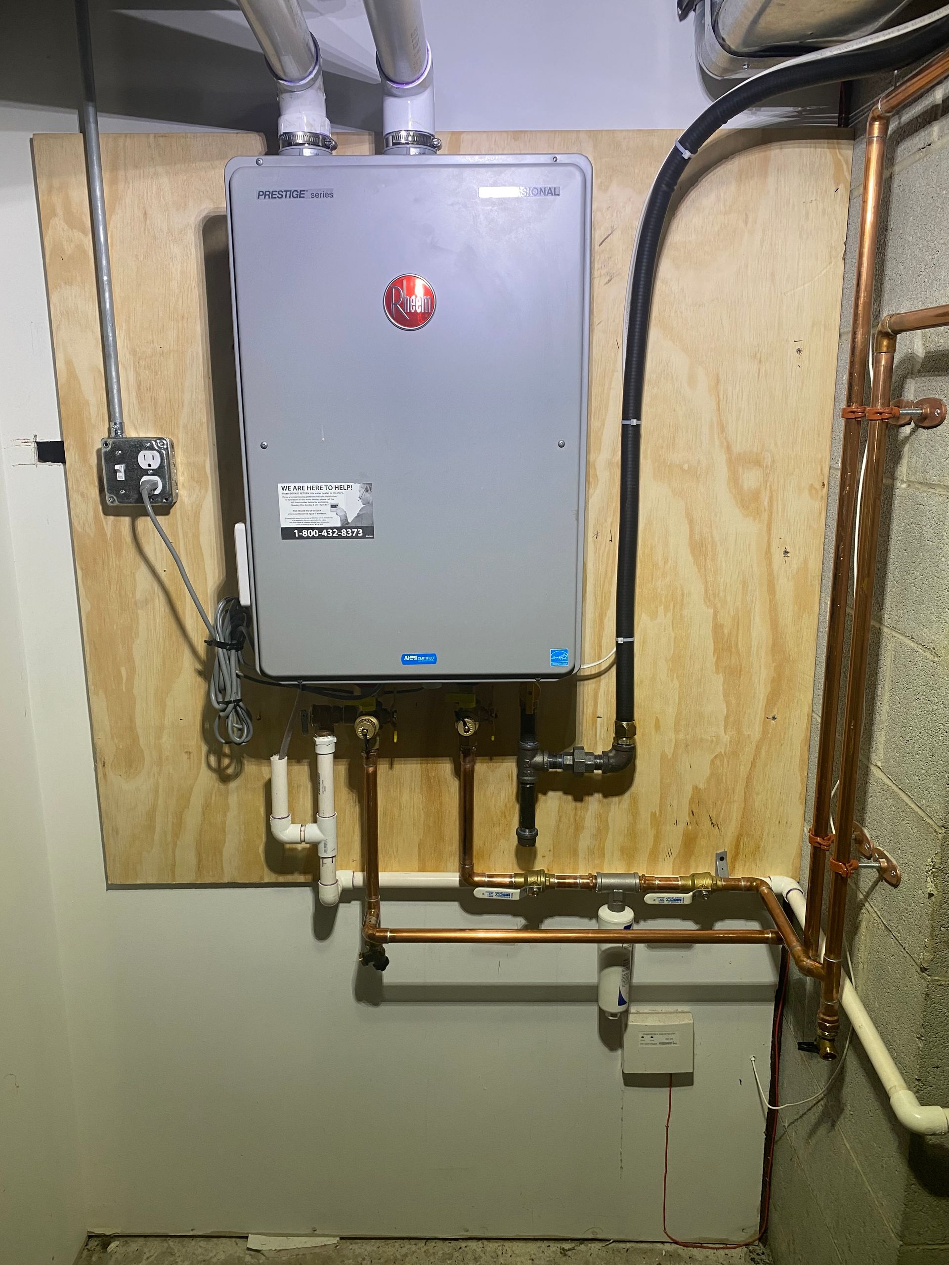 Tankless water heater mounted on plywood in a basement, with copper pipes and venting.