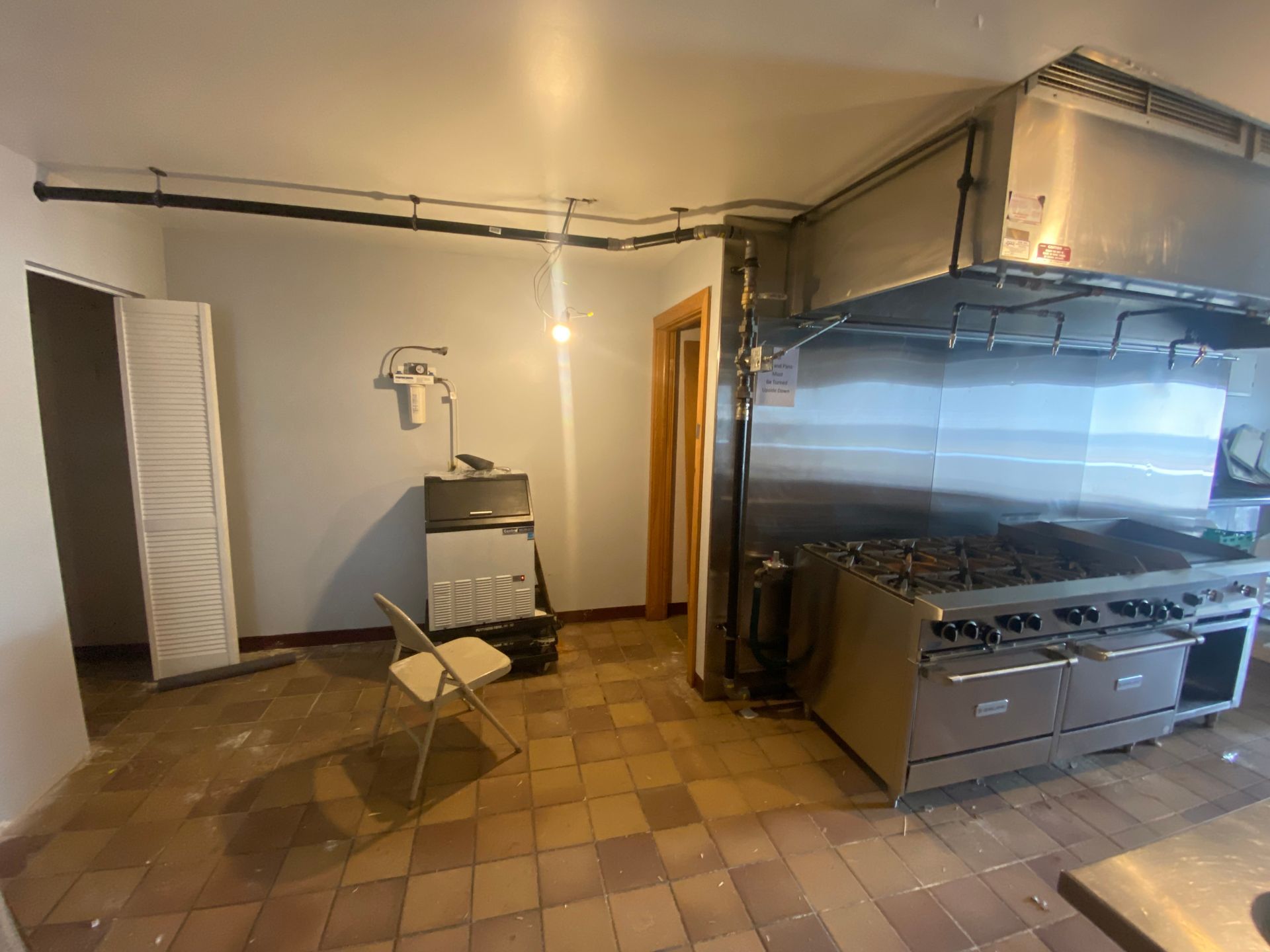 Empty commercial kitchen with stainless steel appliances, tile floor, and exposed pipes.