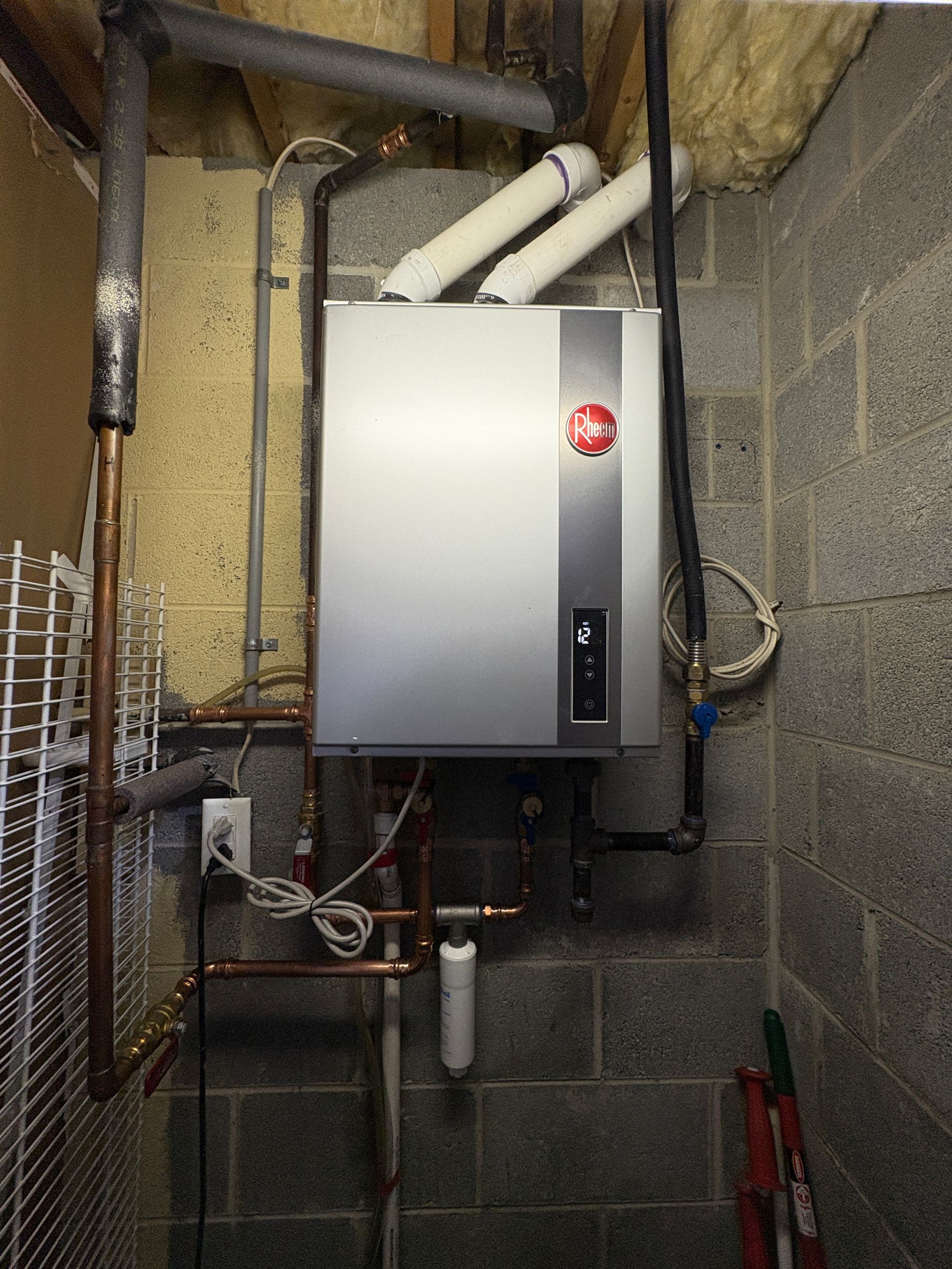 Rheem water heater mounted on a cinder block wall, with pipes and wiring visible.