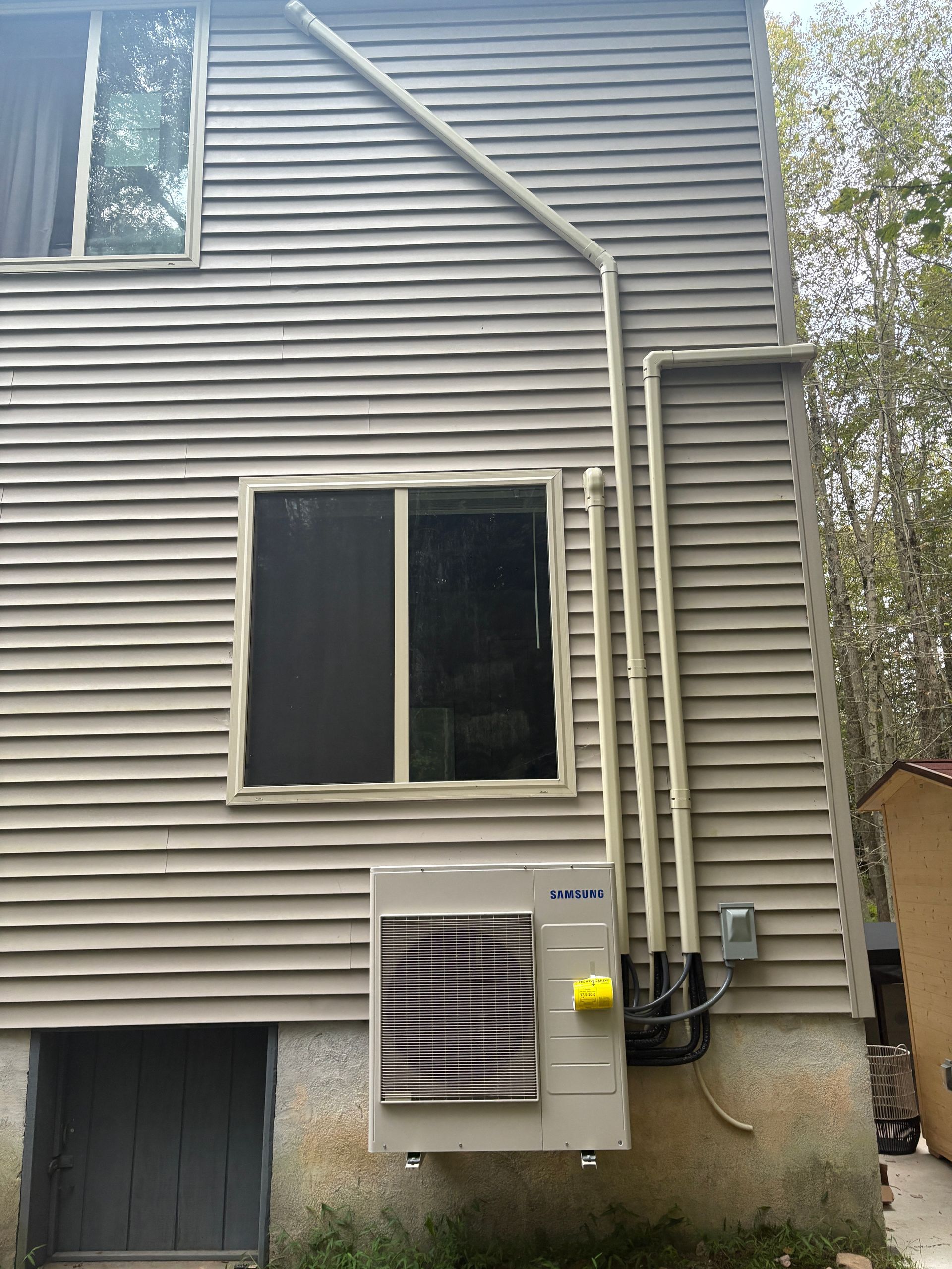 An outdoor HVAC unit mounted on a light brown siding wall with associated piping and electrical box.