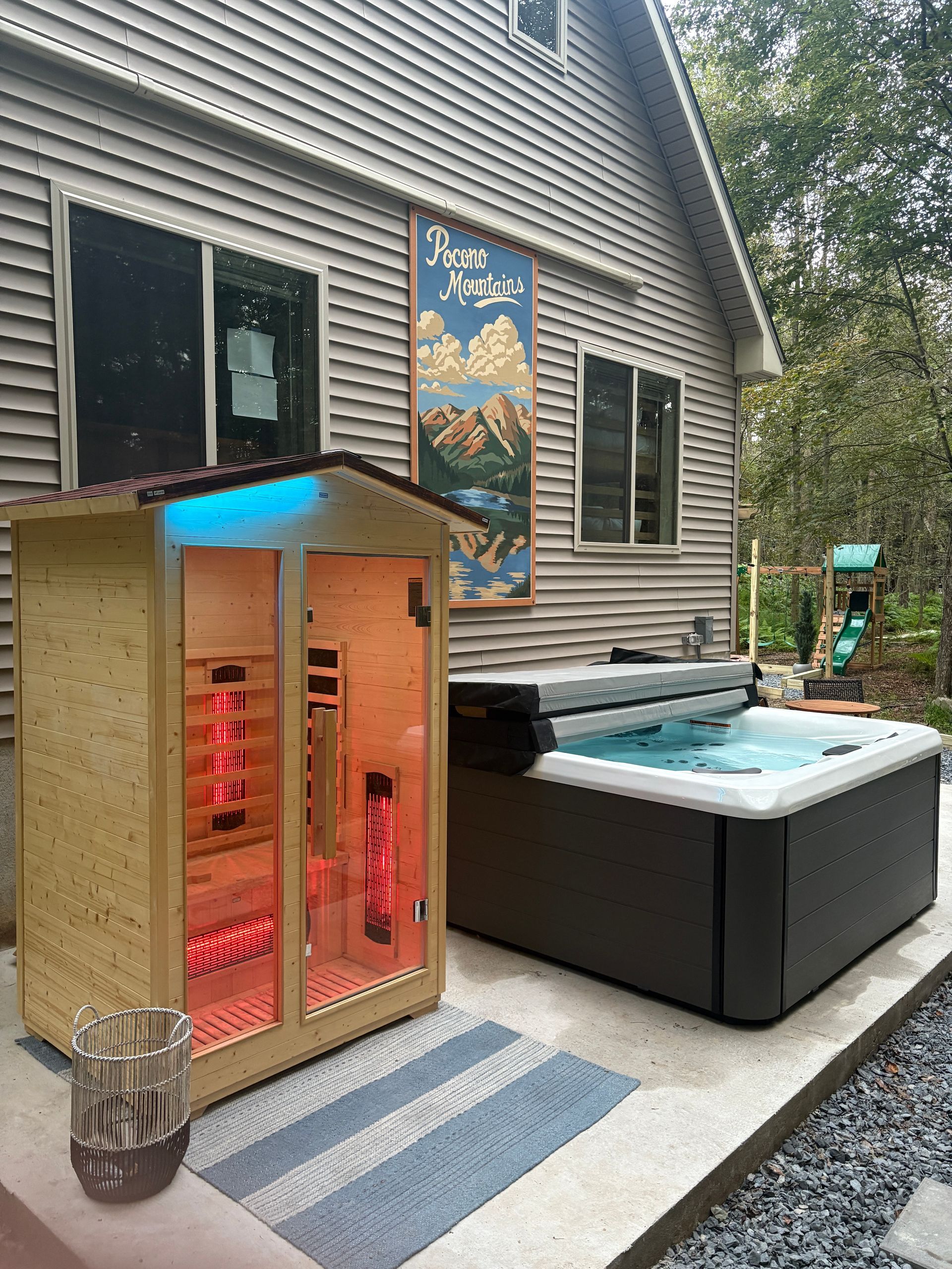 A sauna with red interior lights next to a hot tub outside a house with a colorful wall art.