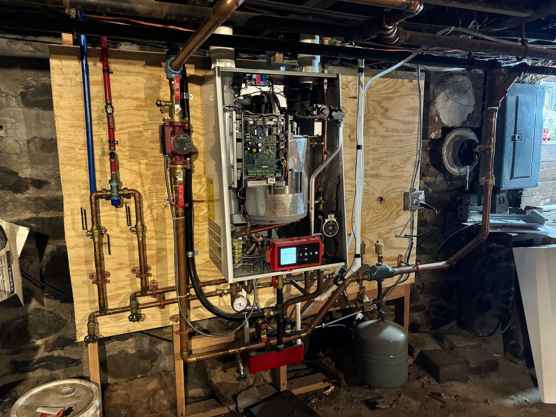 A tankless water heater installed on a plywood board with copper pipes in a basement.