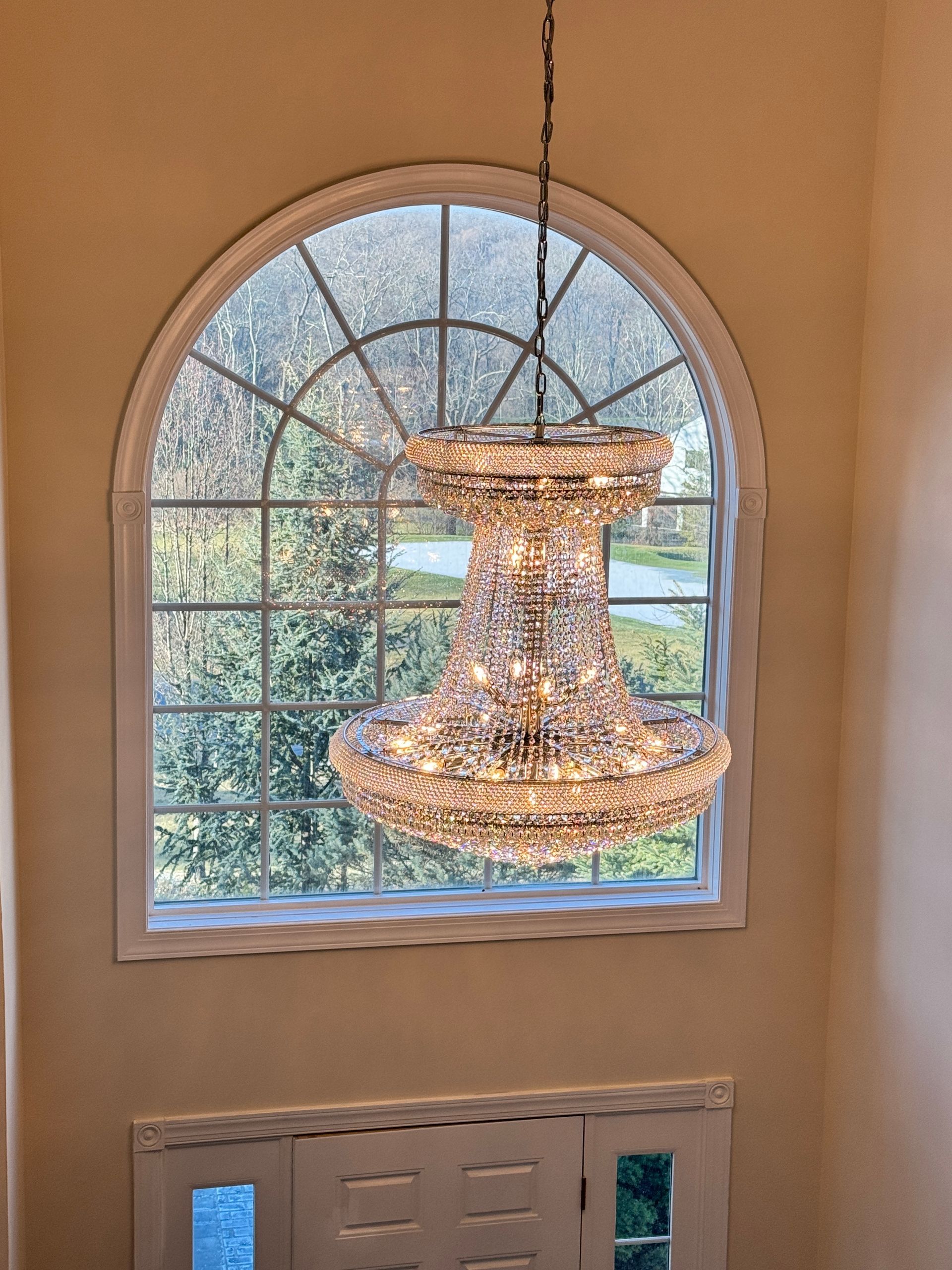 Crystal chandelier hangs in front of arched window. Light beige walls.