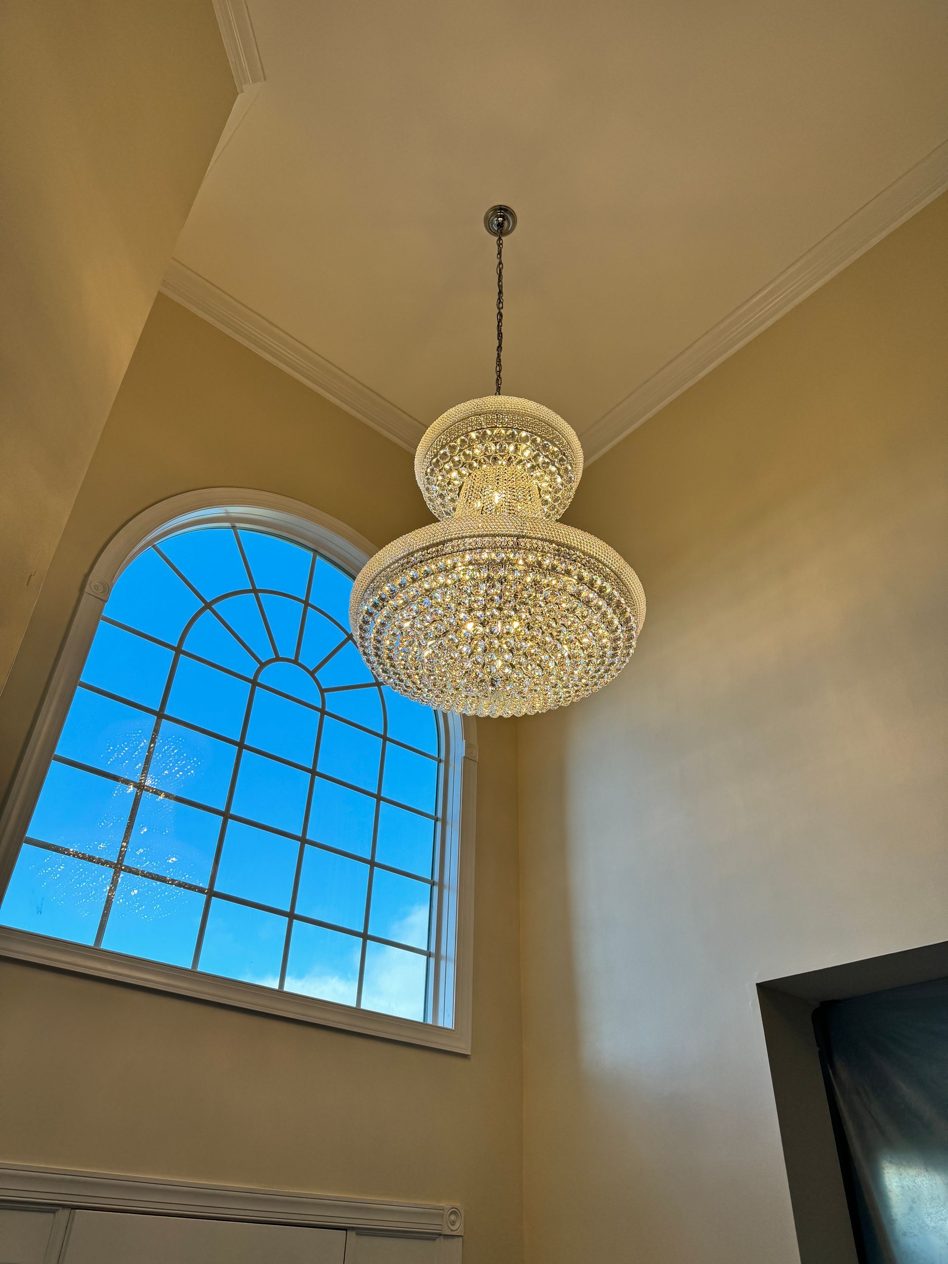 Two-tiered crystal chandelier hangs in front of a large arched window with a blue sky view.