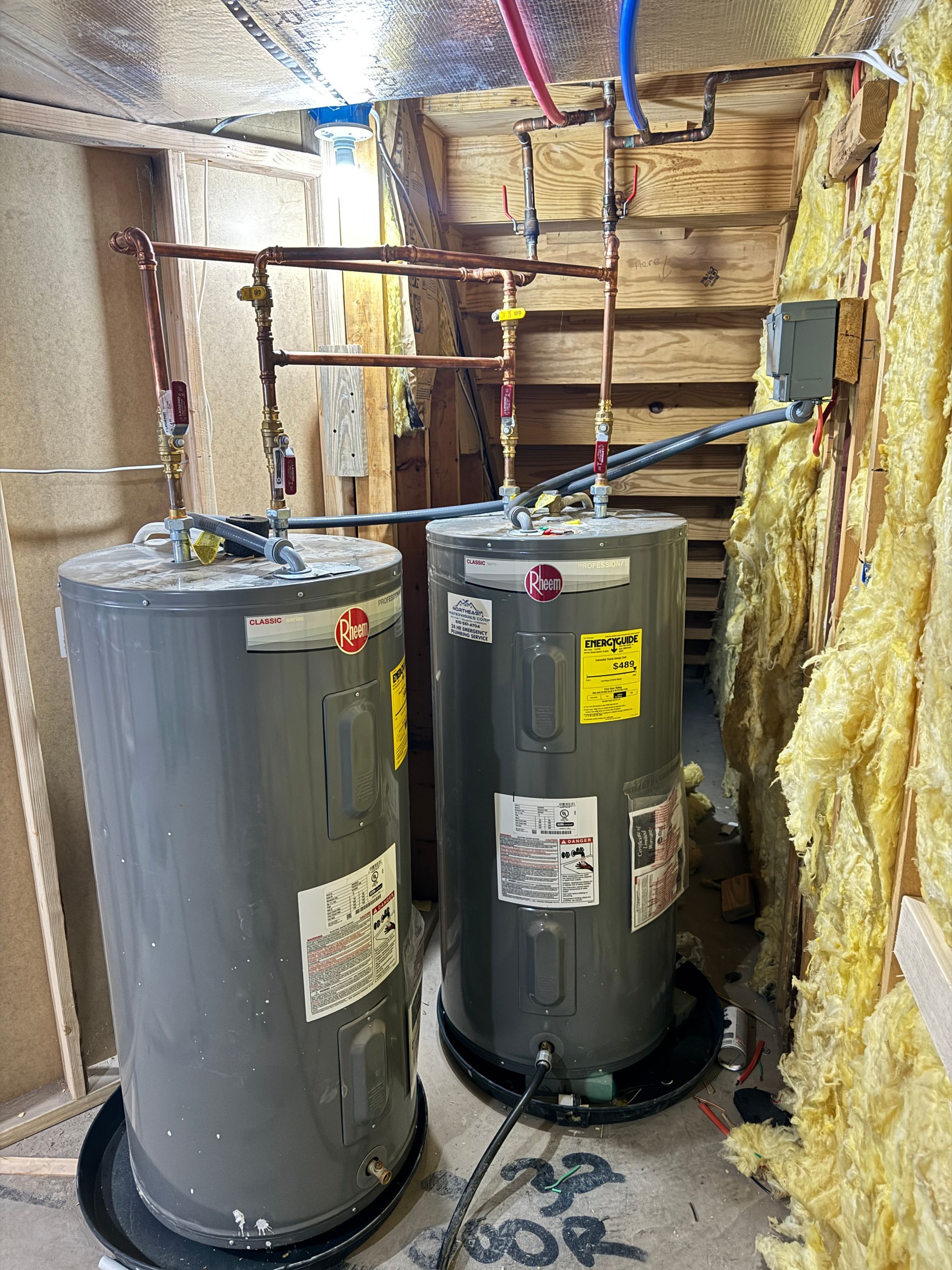 Two gray water heaters with copper pipes, insulation, and electrical connections in a utility space.