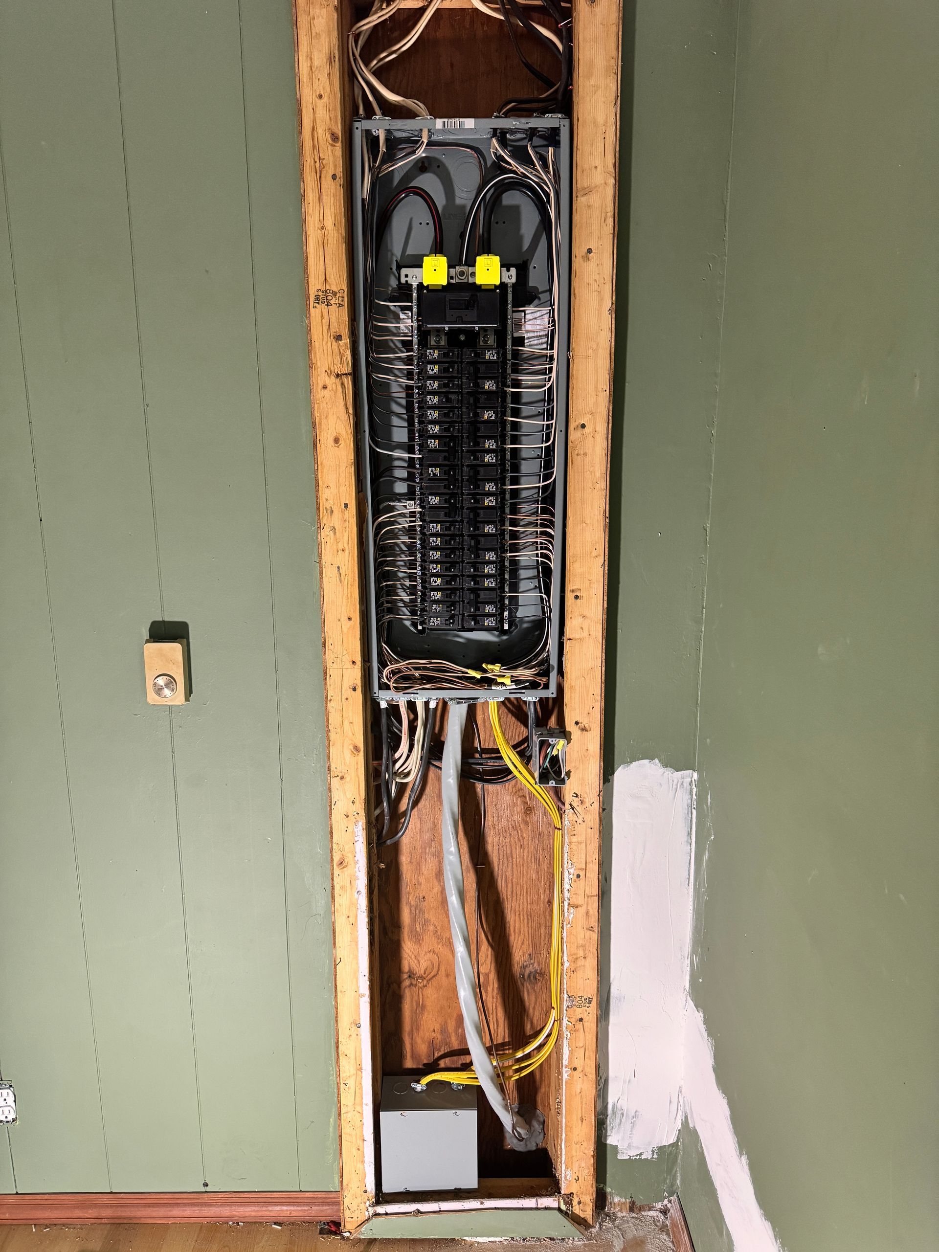 Electrical panel installed in a wall opening, wires exposed, on green painted wall.