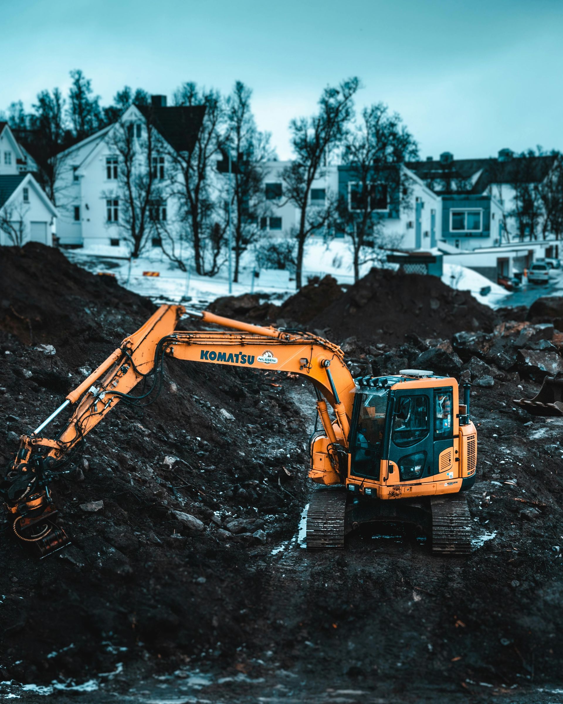 Yellow excavator on a mound of dark soil, houses in the background under a cloudy sky.